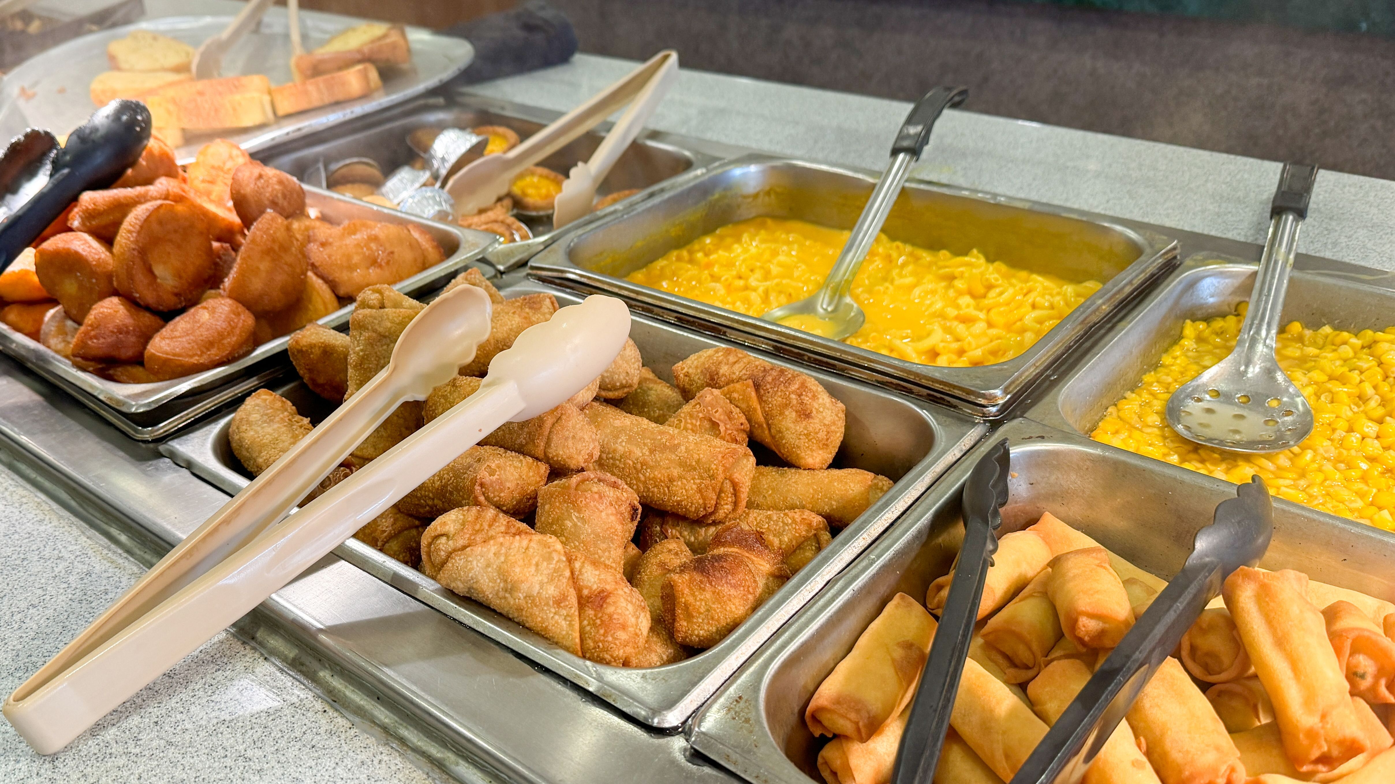 a buffet-style food setup with trays of various fried items, such as spring rolls and breaded snacks, alongside macaroni and cheese, and corn a buffet-style food setup with trays of various fried items, such as spring rolls and breaded snacks, alongside macaroni and cheese, and corn