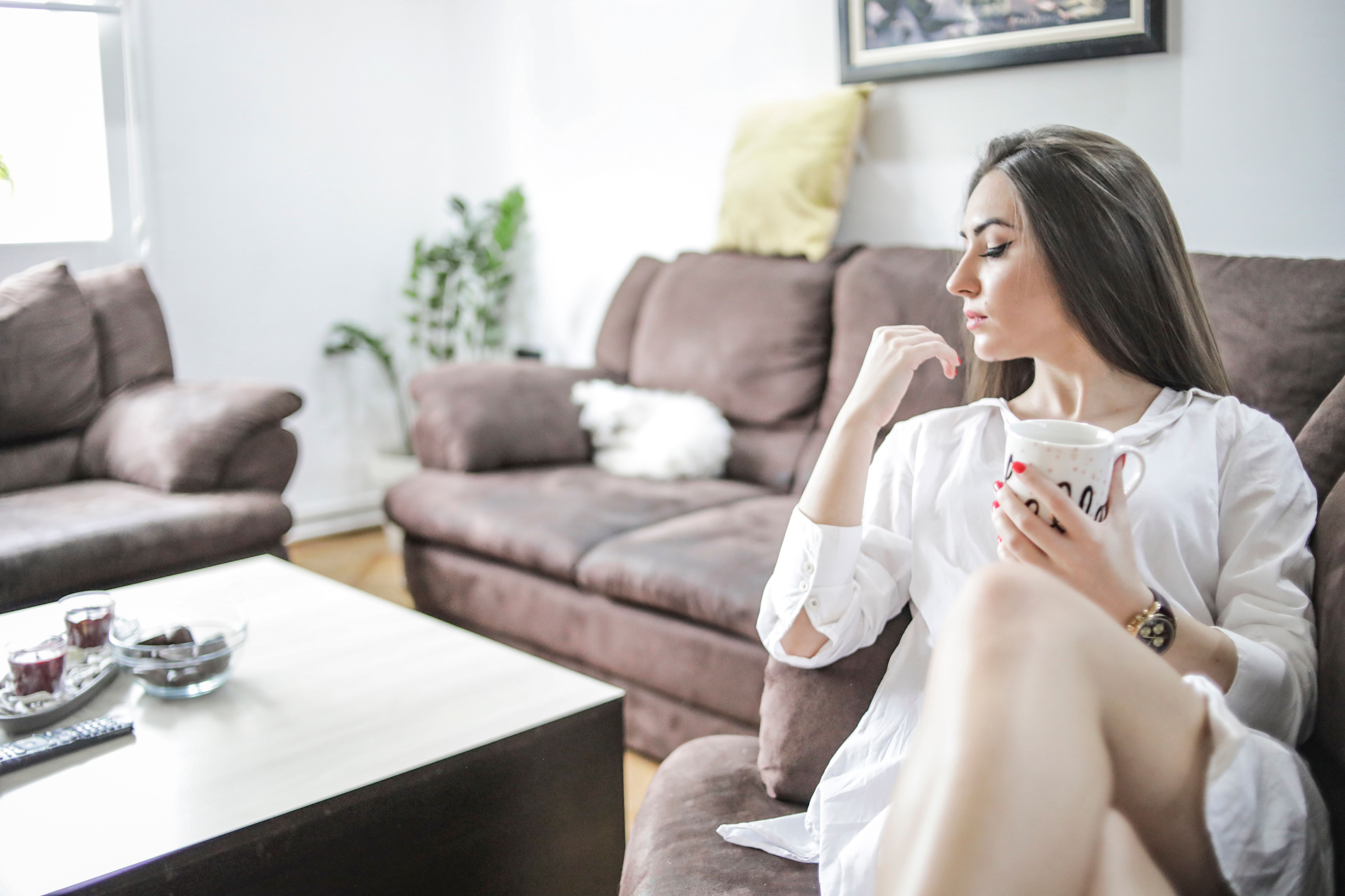 A beautiful young woman in a men's shirt is resting in her home, sitting on a sofa in the living room, enjoying sexuality and a free weekend.