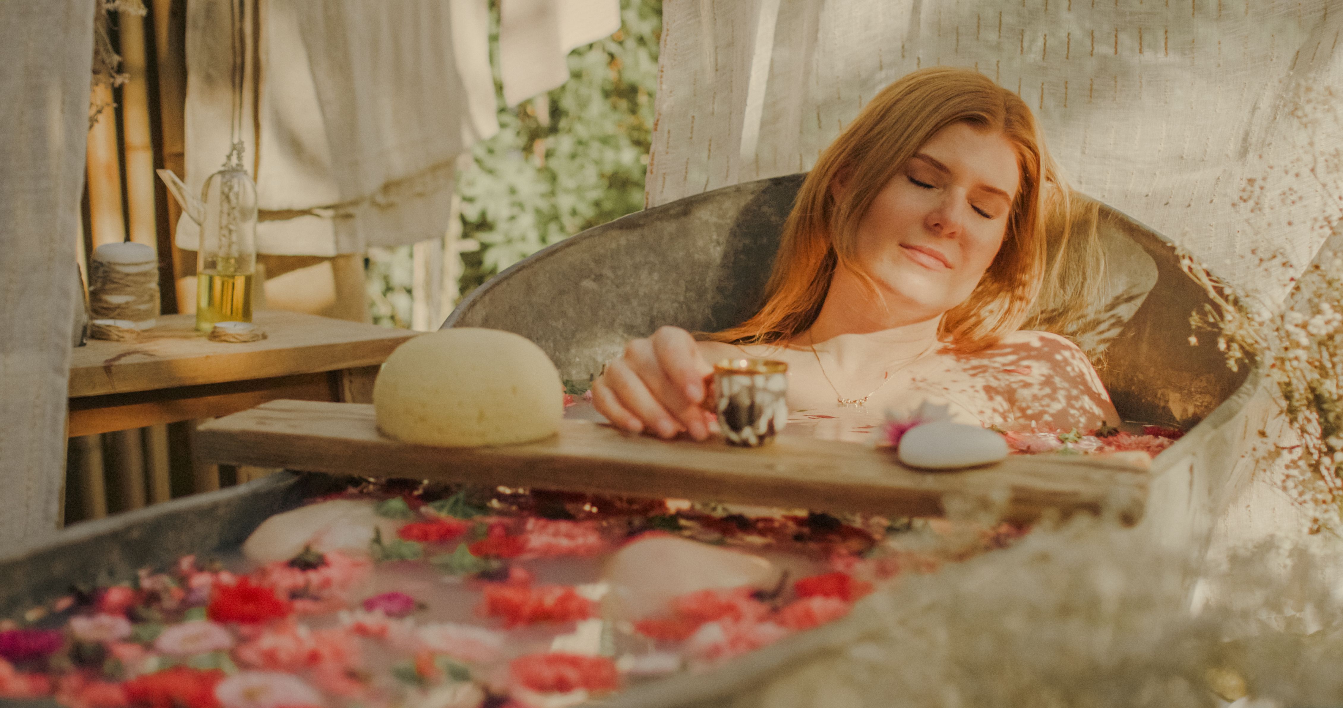 A red-haired woman drinks coffee in a bathtub full of flowers. The bathtub is in the garden.
