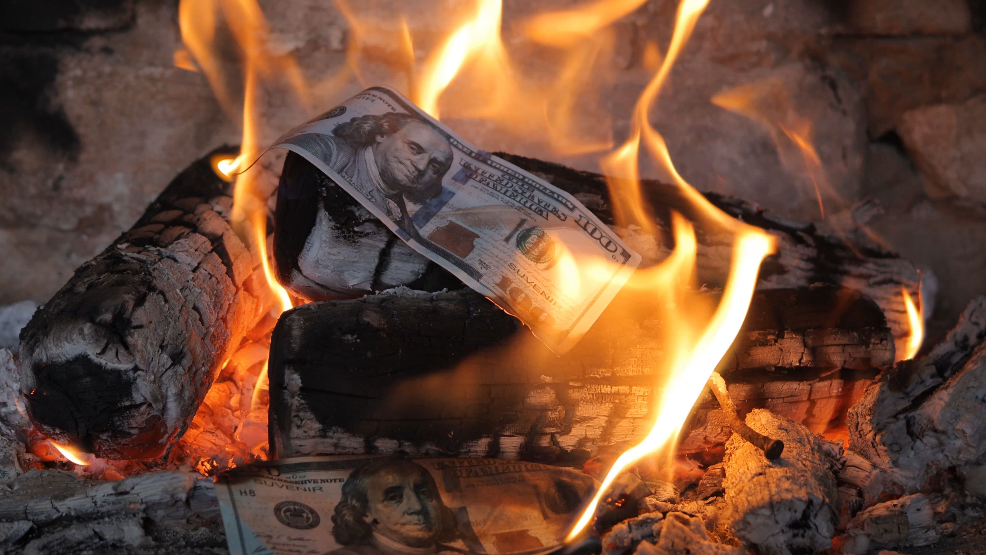 Symbolic image of burning US dollar bills amidst fiery logs in a fireplace, representing financial loss, economic crisis, or wasted wealth. Bright flames consume the currency.