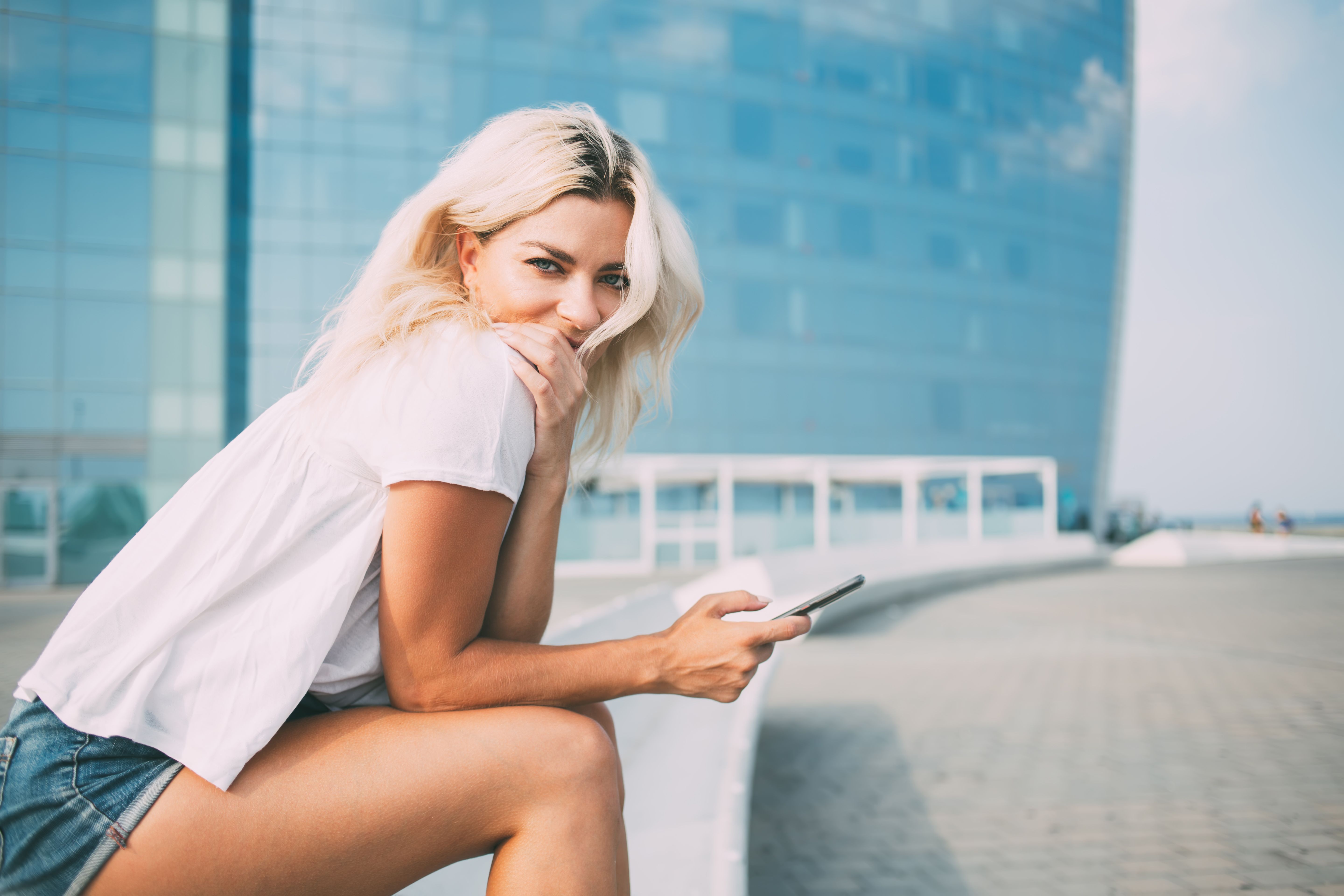 Portrait of charming hipster girl in casual outfit holding modern cellular device and looking at camera during leisure time in city, millennial female user with smartphone posing at urban setting
