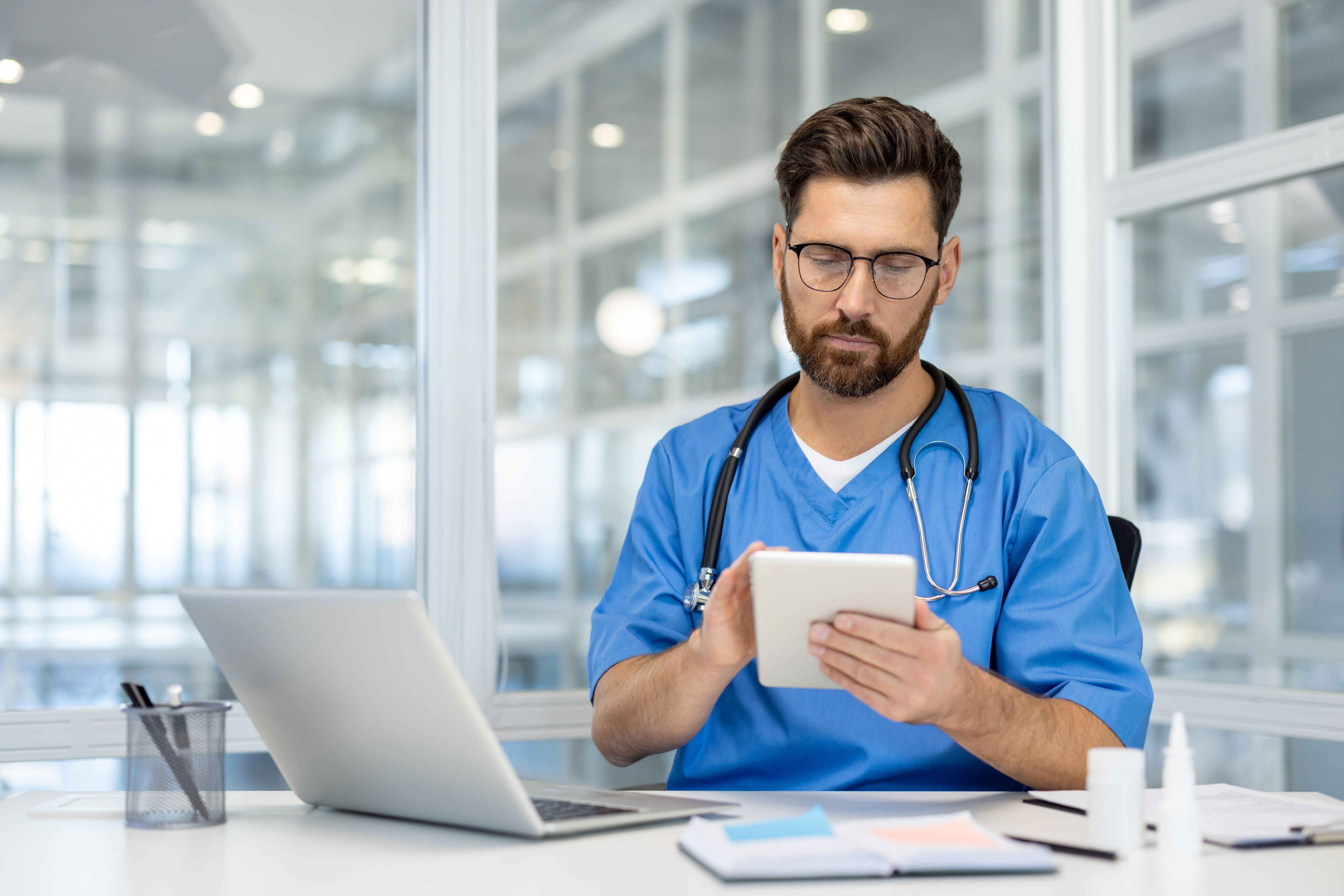 Professional doctor in blue scrubs using a digital tablet at modern medical office. Focused healthcare professional accessing patient records online to optimize healthcare delivery.