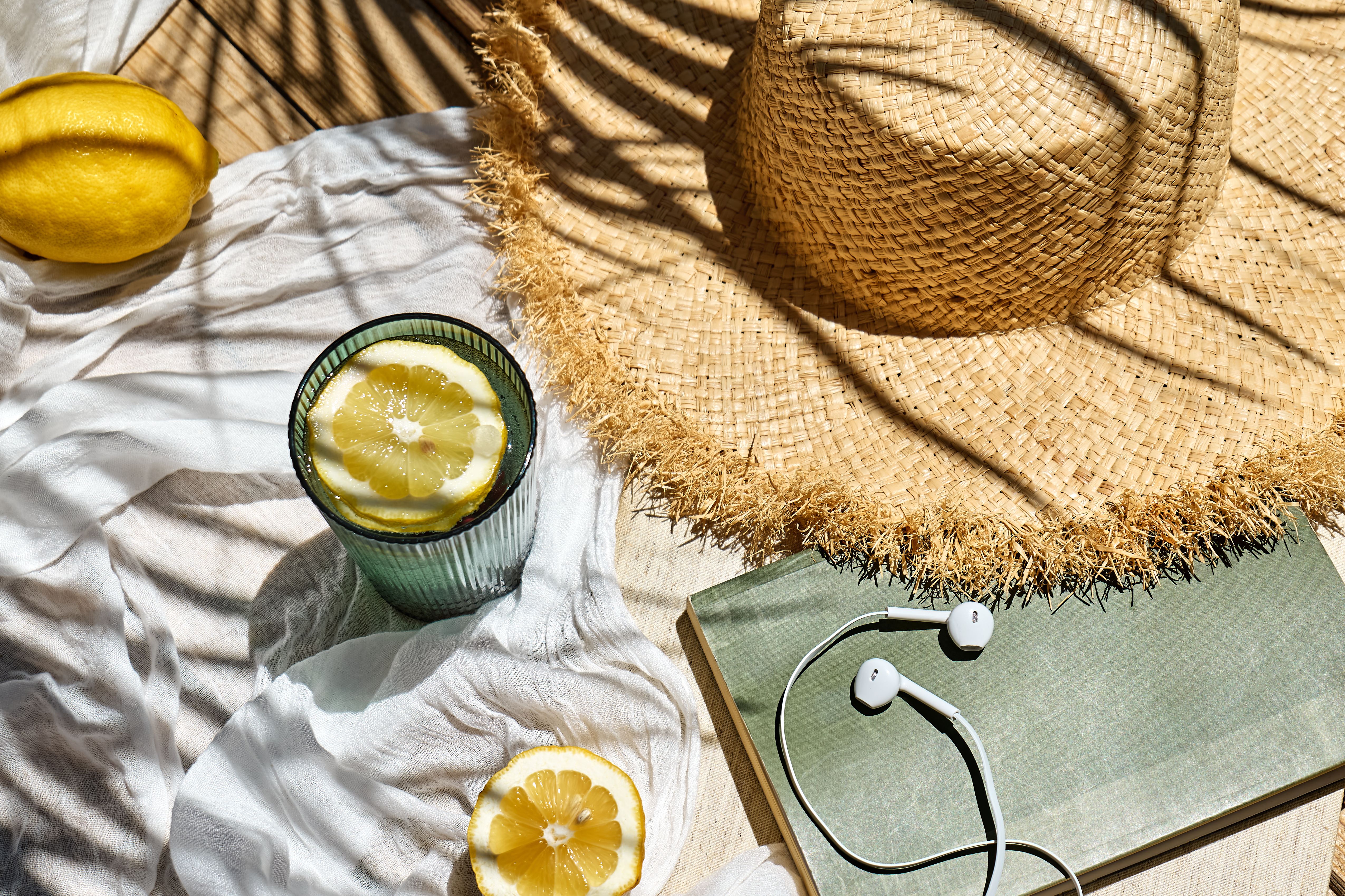 Audiobook or podcast during summer holidays. Headphones, book, rifresh drink, sunglasses and hat on white beach blanket. Summertime mood. Audiobook or podcast during summer holidays. Headphones, book, rifresh drink, sunglasses and hat on white beach blanket. Summertime mood.