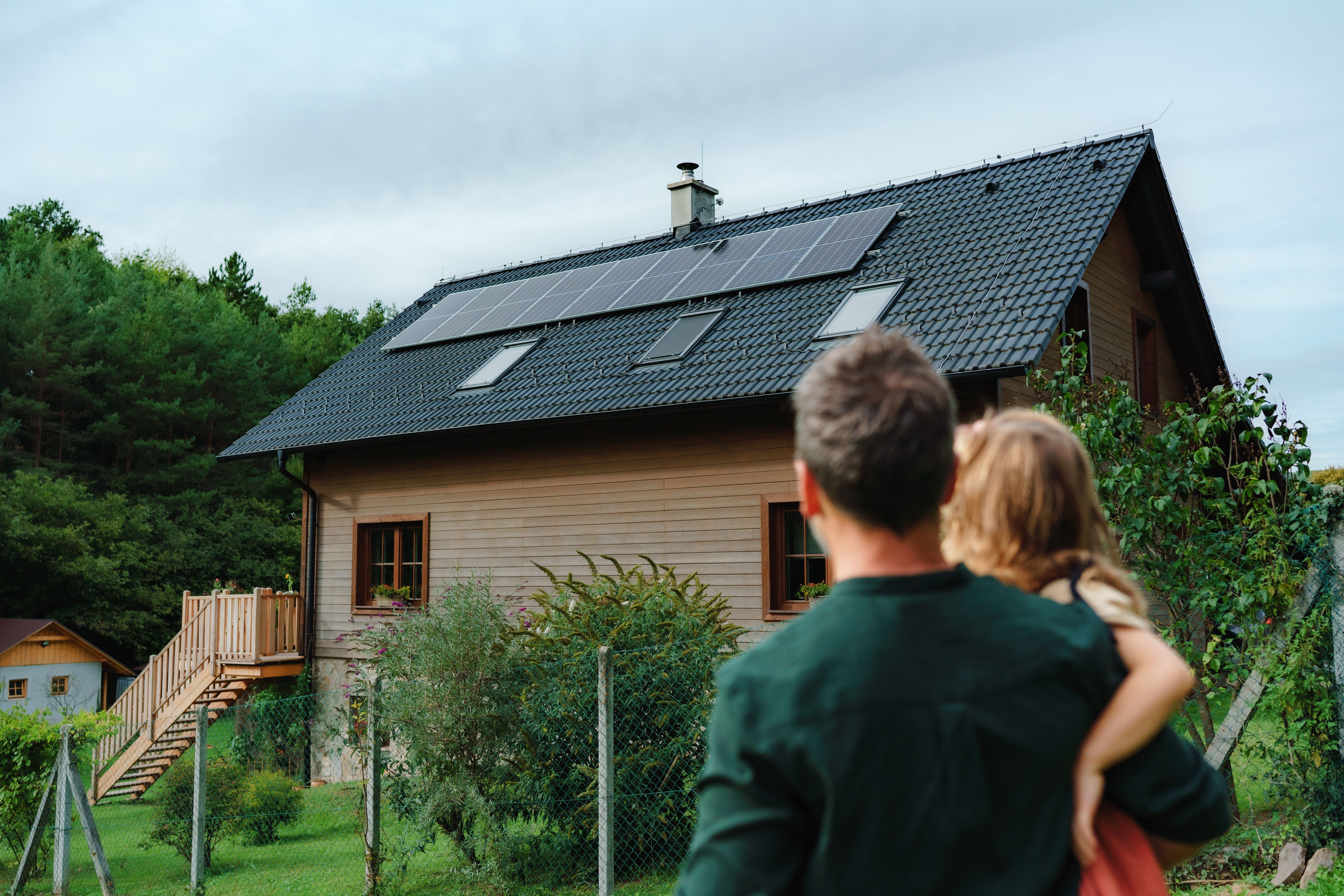 Rear view of dad holding her little girl in arms and looking at their house with installed solar panels. Alternative energy, saving resources and sustainable lifestyle concept.