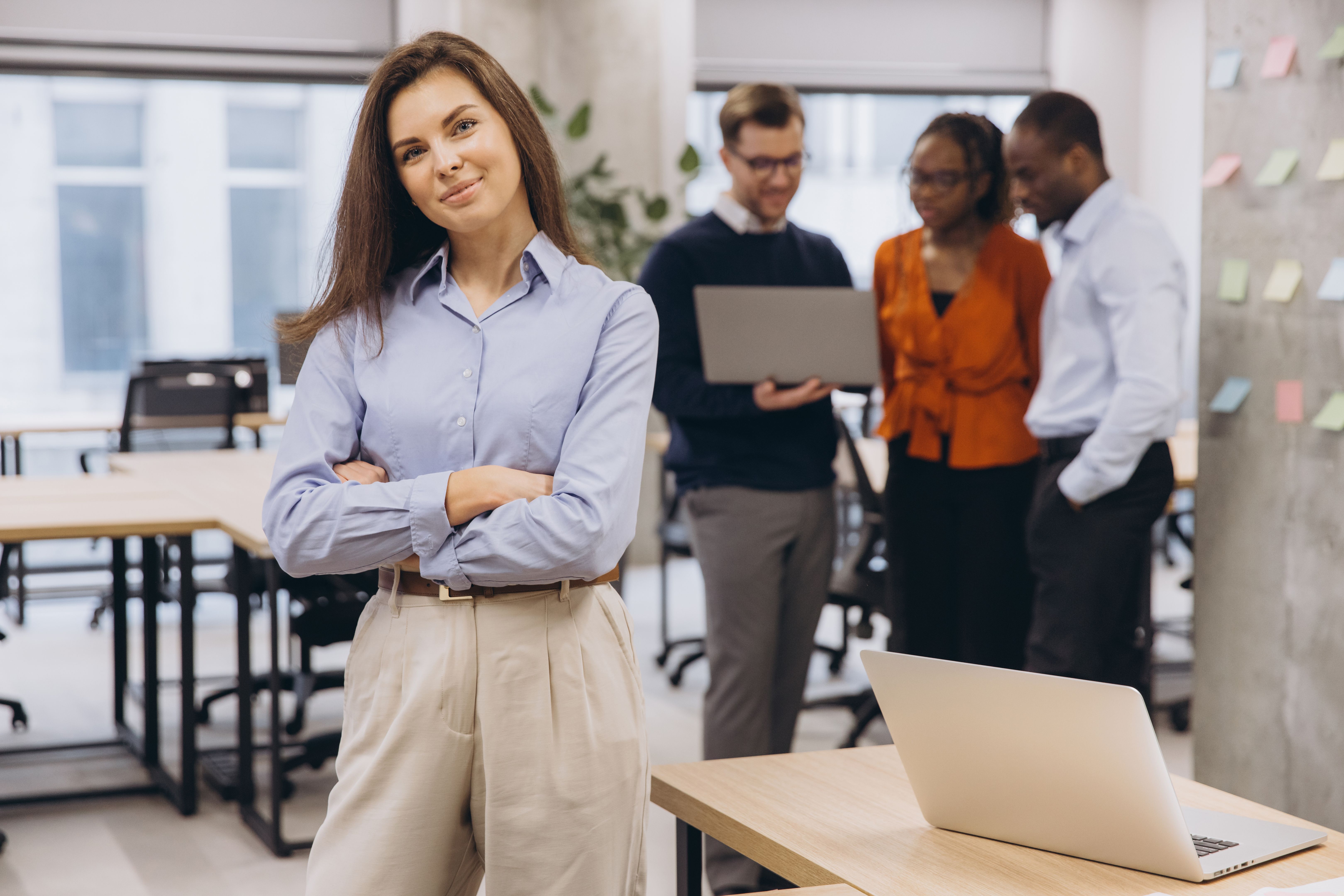 Portrait of a businesswoman standing with arms crossed in a modern office, her colleagues collaborating in the background, showcasing teamwork and leadership