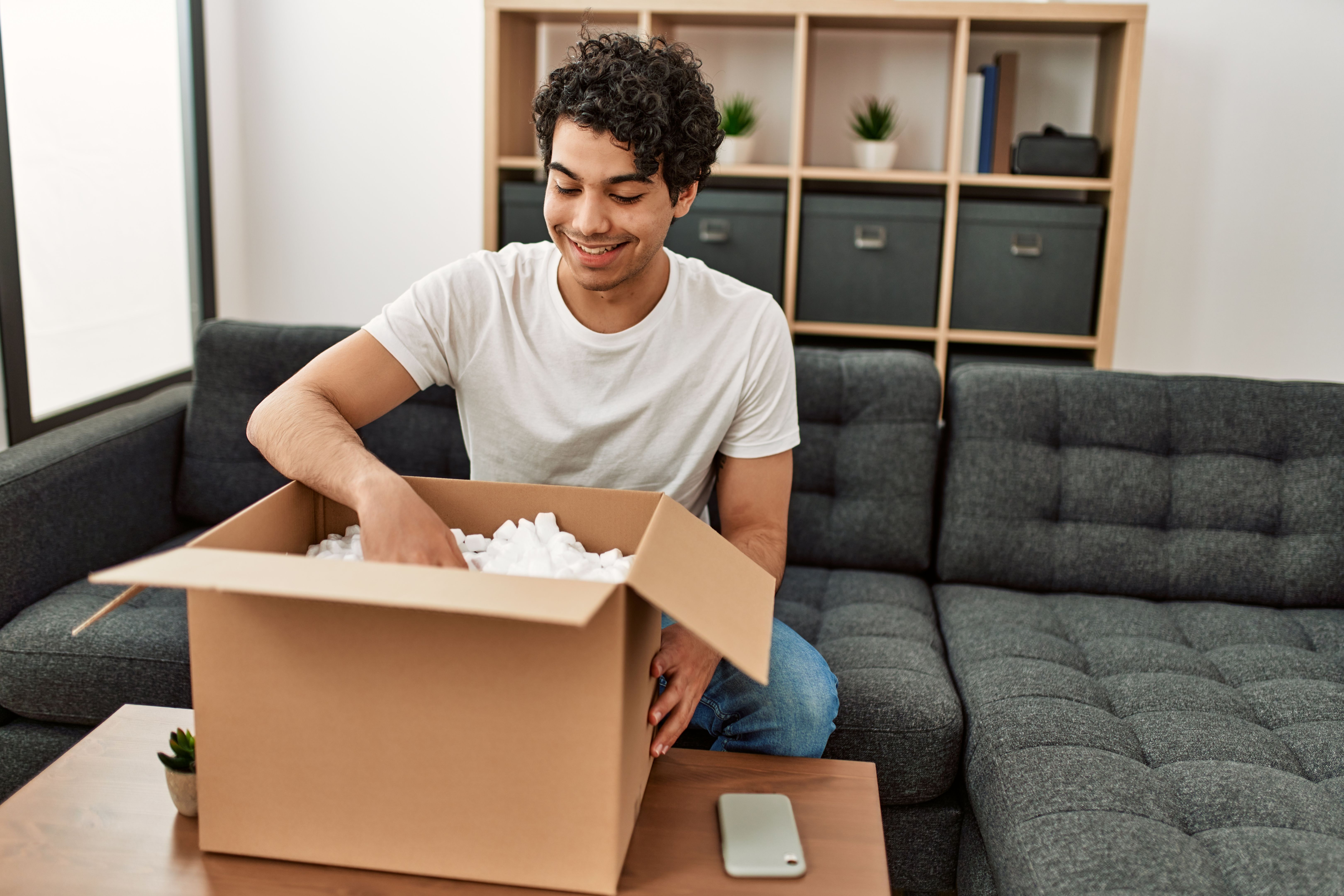 Young hispanic man unboxing cardboard box sitting on the sofa at home.