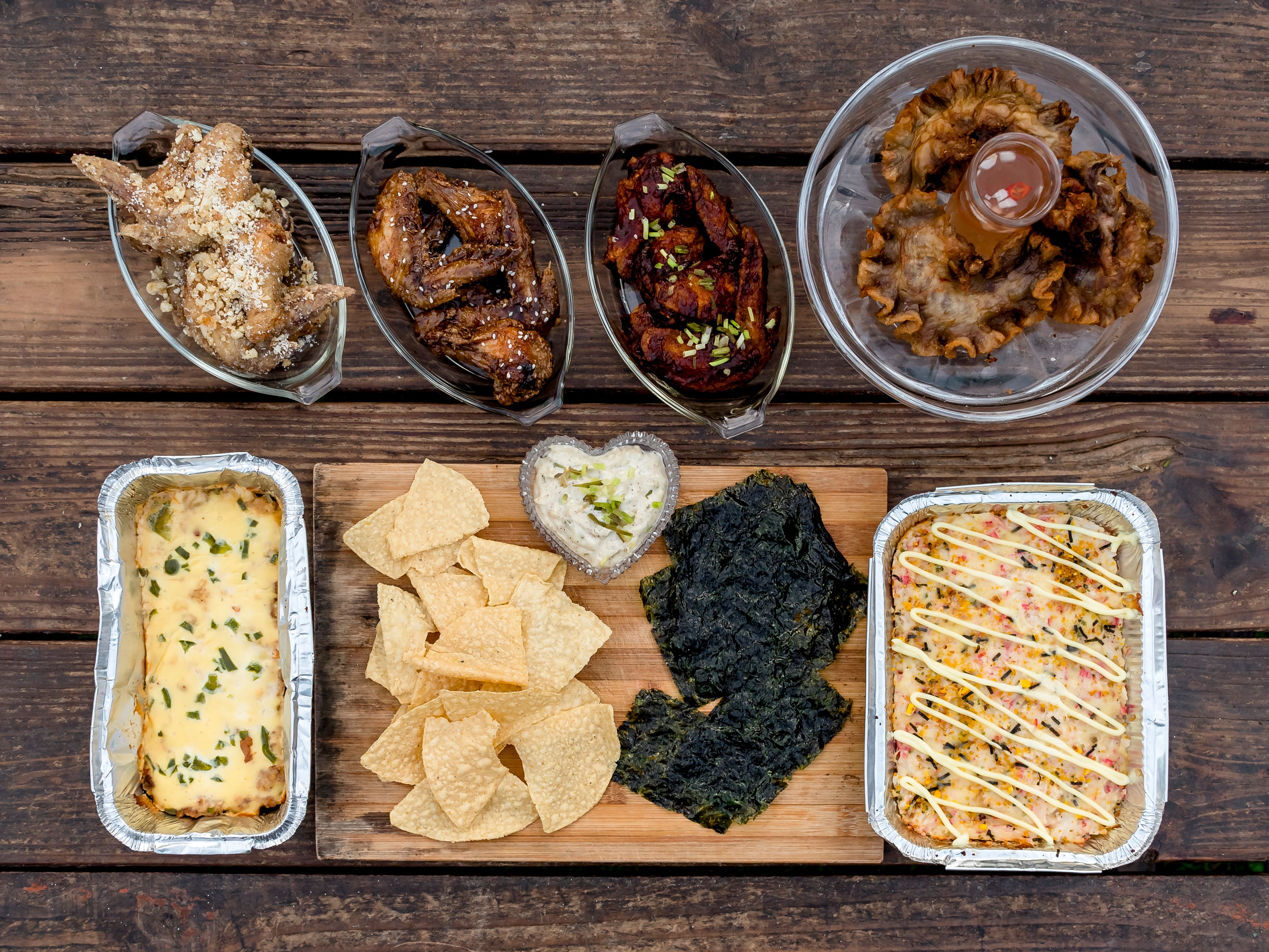 An assortment of food items on a wooden bench: Chicken wings, Chicharon Bulaklak, Baked Nachos and Baked Sushi. An assortment of food items on a wooden bench: Chicken wings, Chicharon Bulaklak, Baked Nachos and Baked Sushi.