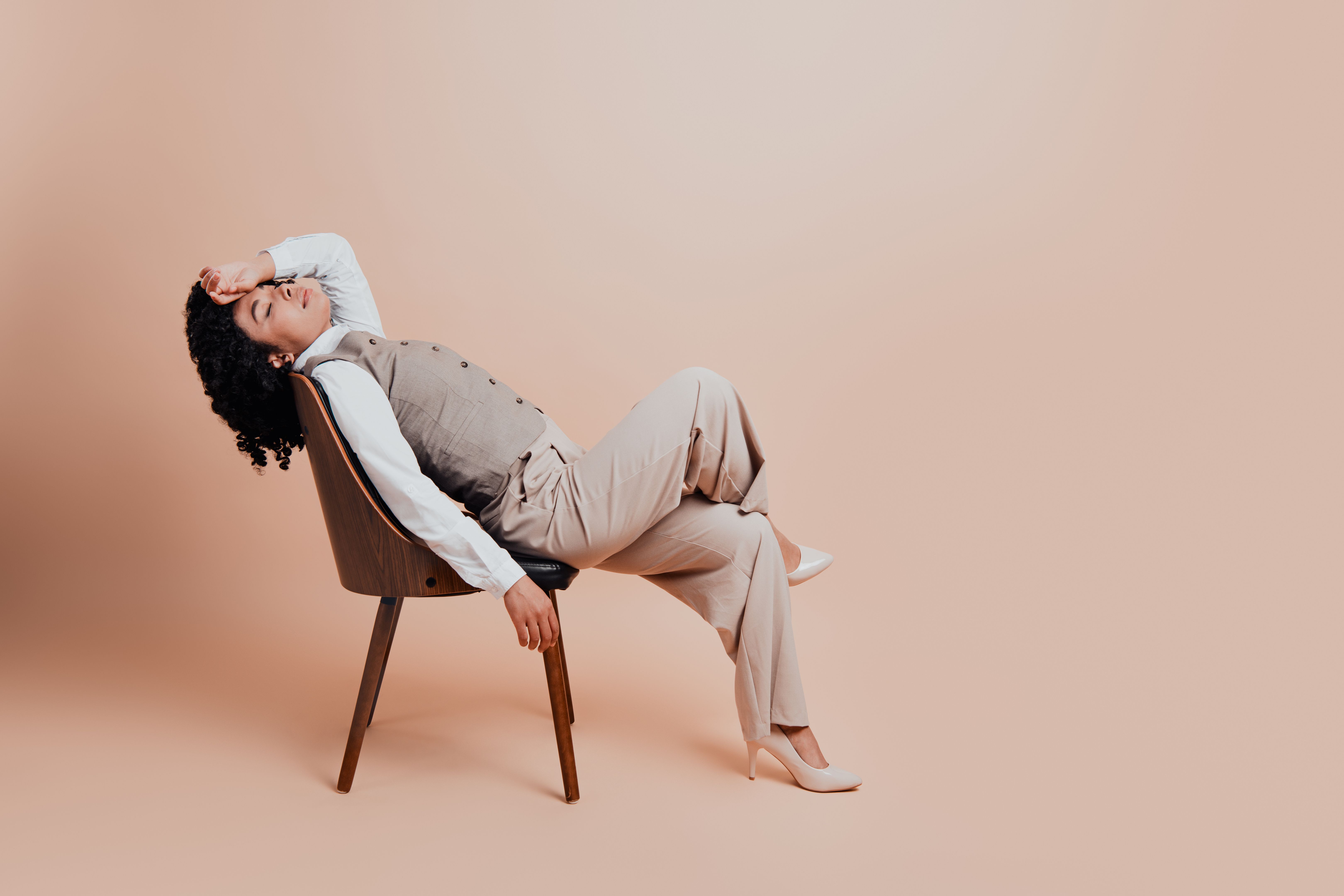 Professional woman wearing formal attire sitting on a wooden chair in a stylish pose against a beige background