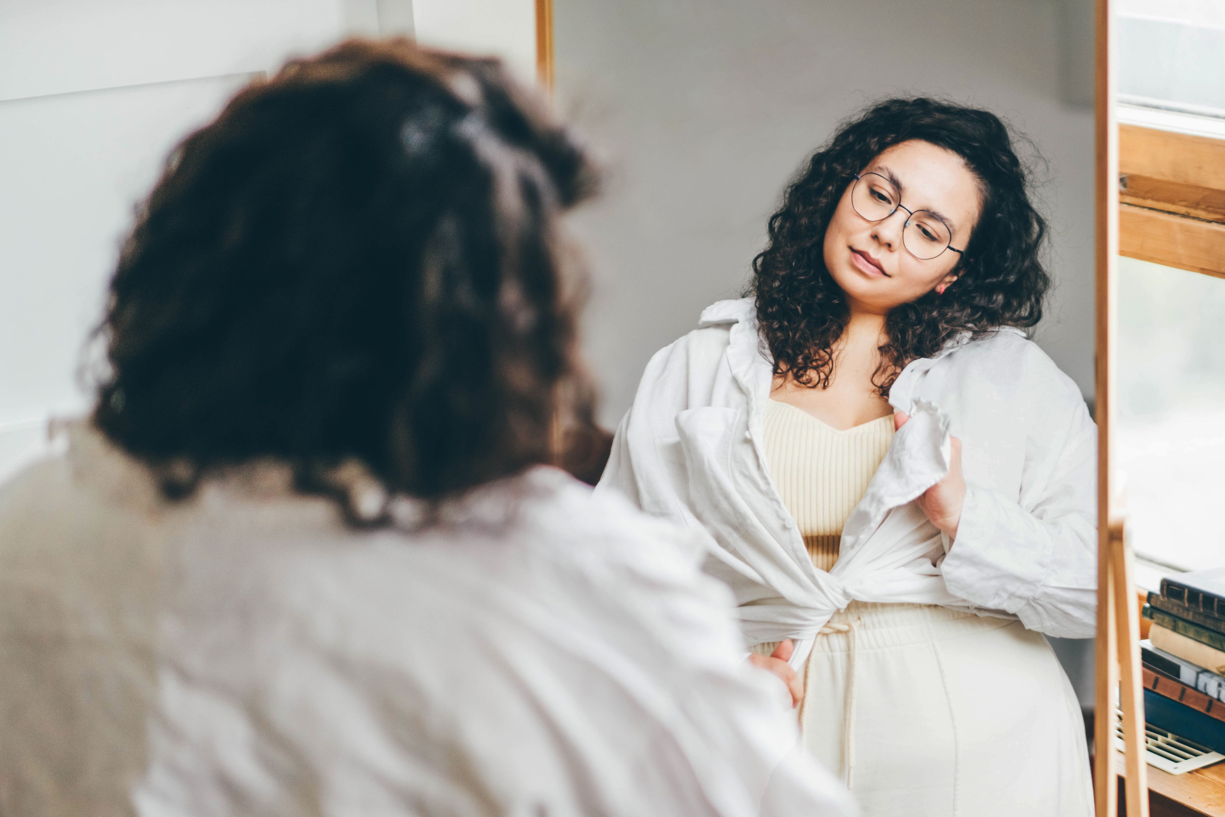 Curly haired overweight young woman in glasses ties white shirt and admires choice standing in front of large mirror in stylish room reflection view