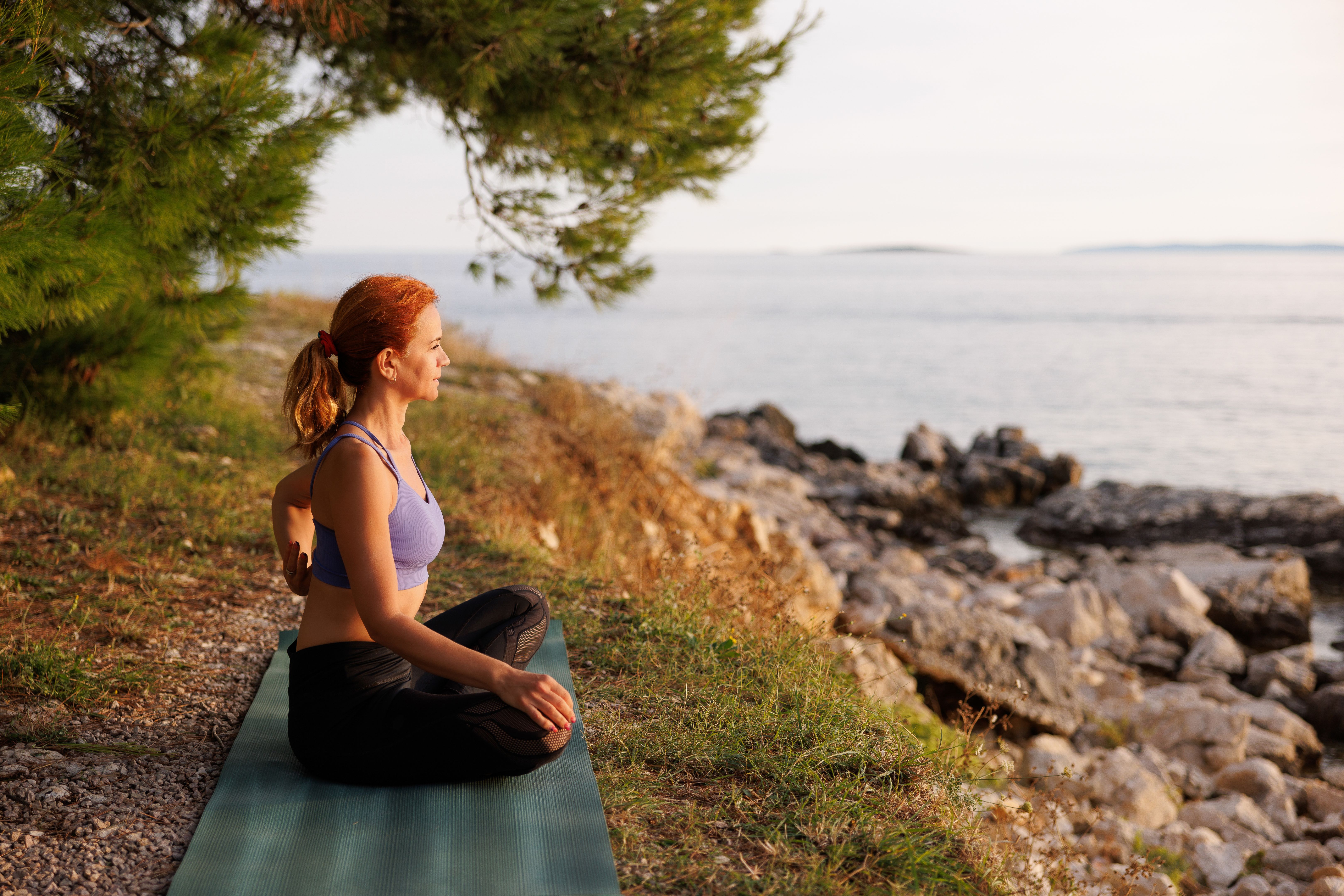 Healthy woman sitting cross-legged on exercise mat and relaxing in the nature by the rocky coastline Healthy woman sitting cross-legged on exercise mat and relaxing in the nature by the rocky coastline