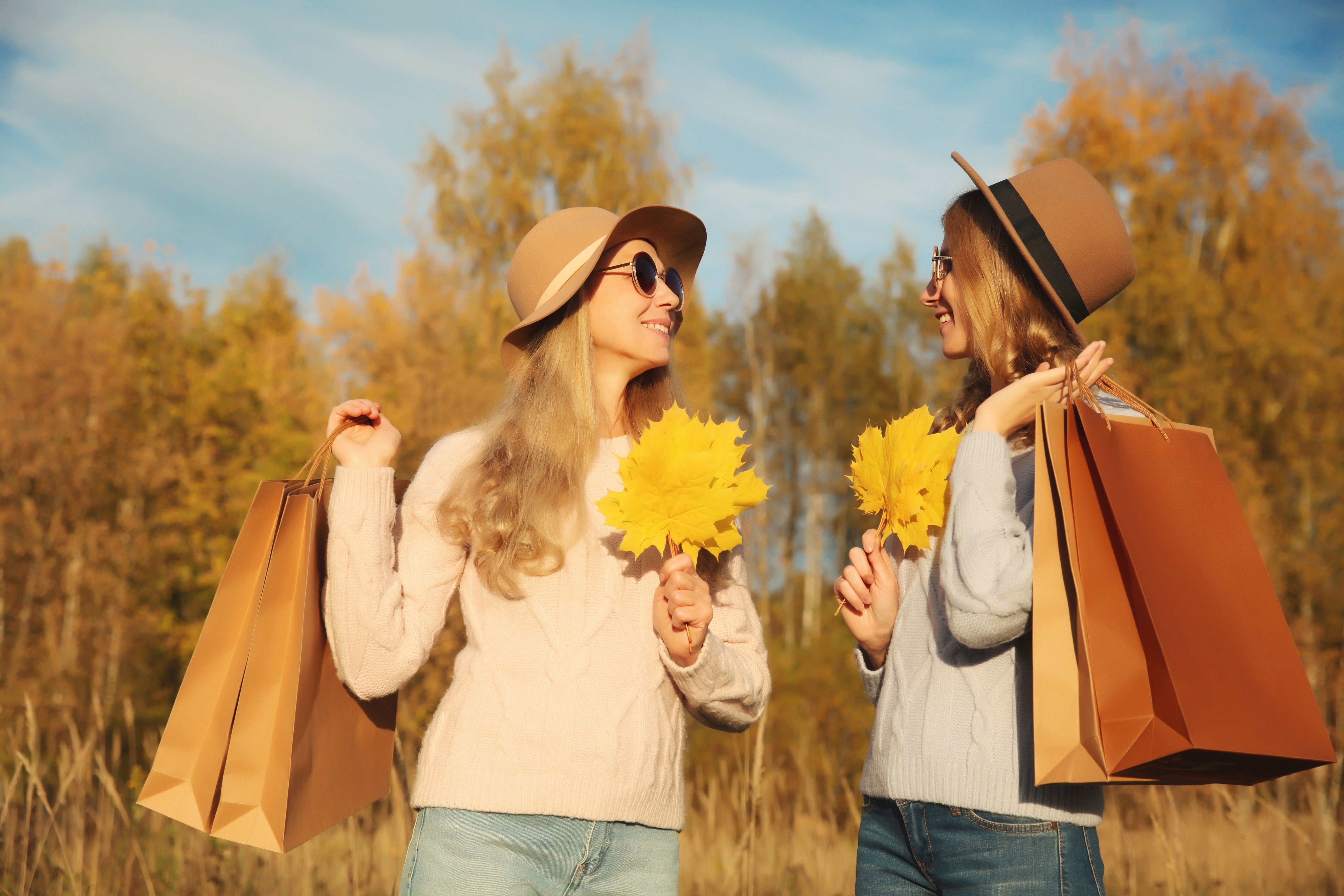 Happy two women friends with autumn yellow leaves, shopping bags, stylish girlfriends smile in park