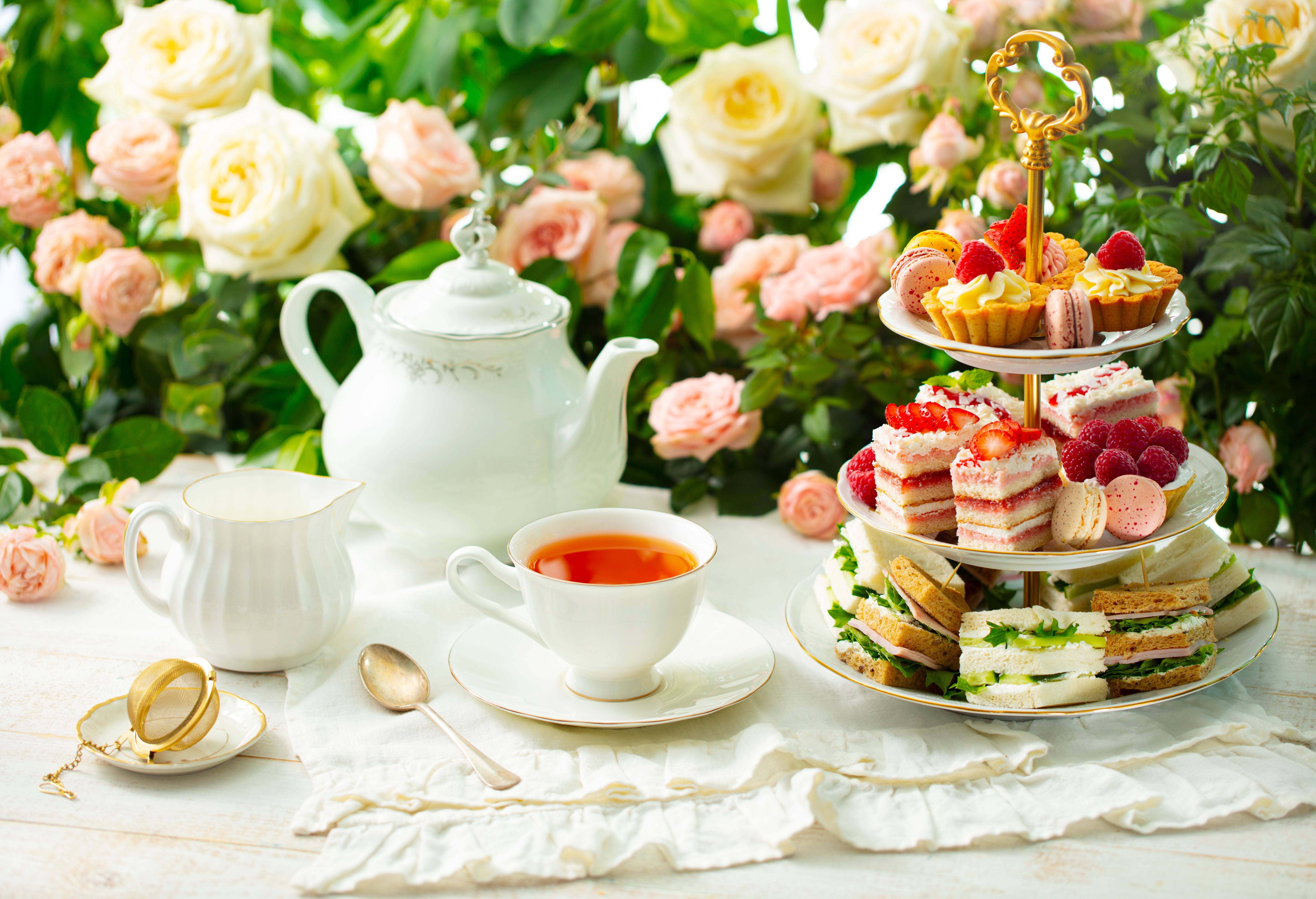 Afternoon tea table. Assortment of cakes, sweets and sandwiches. Rose garden background.