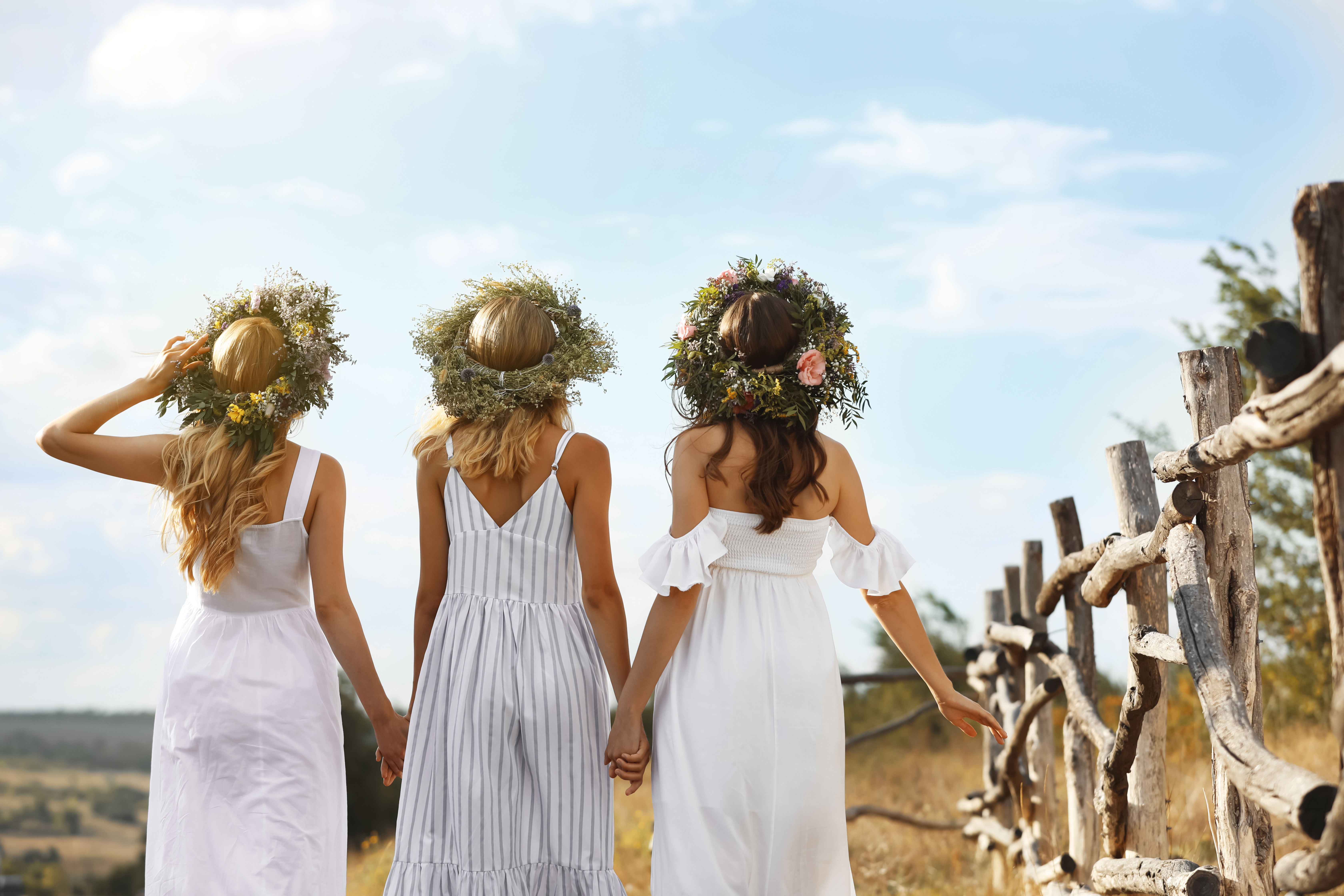 Young women wearing wreaths made of beautiful flowers outdoors on sunny day, back view Young women wearing wreaths made of beautiful flowers outdoors on sunny day, back view