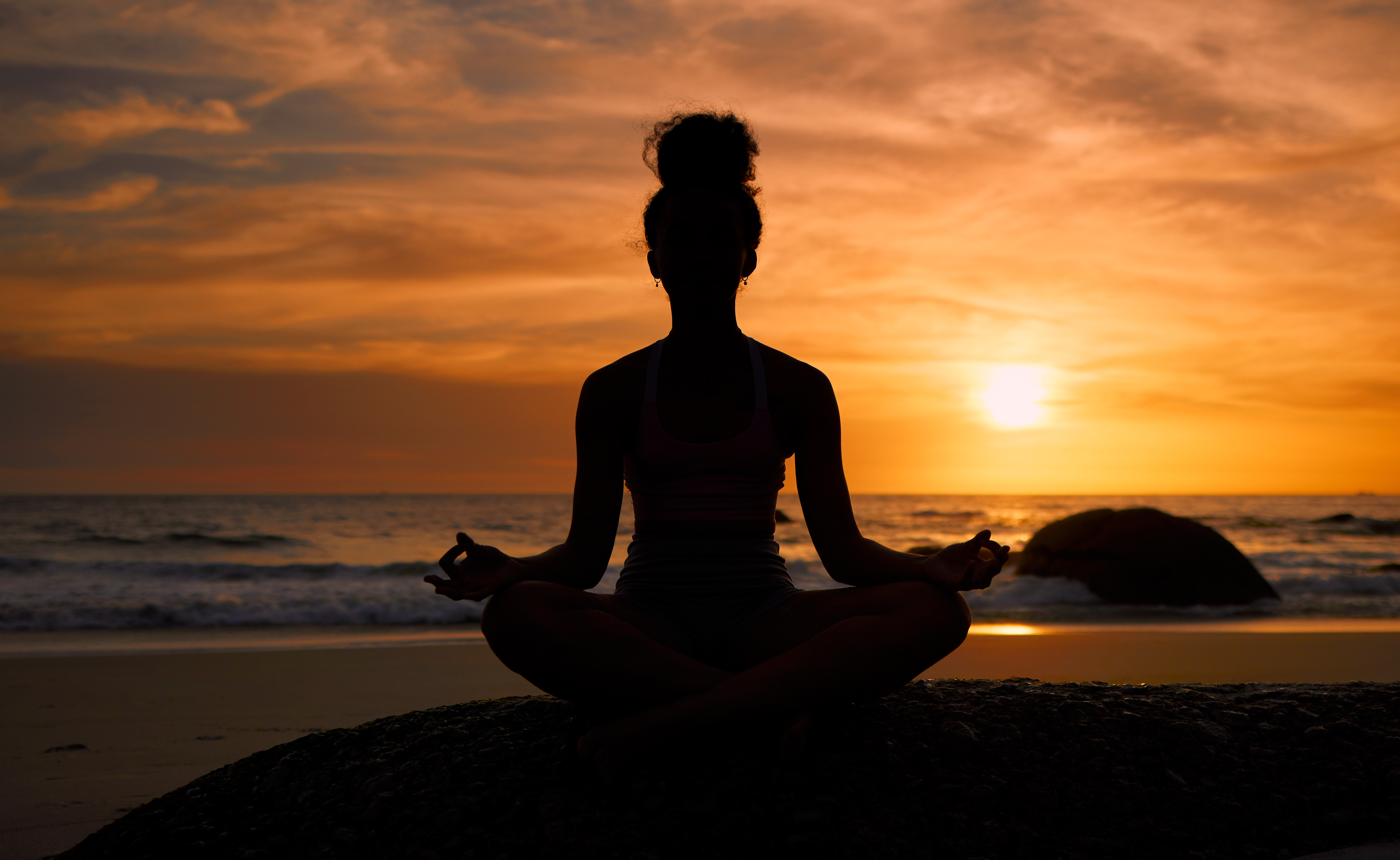 Sunset, beach and silhouette of a woman in a lotus pose while doing a yoga exercise by the sea. Peace, zen and shadow of a calm female doing meditation or pilates workout outdoor at dusk by the ocean