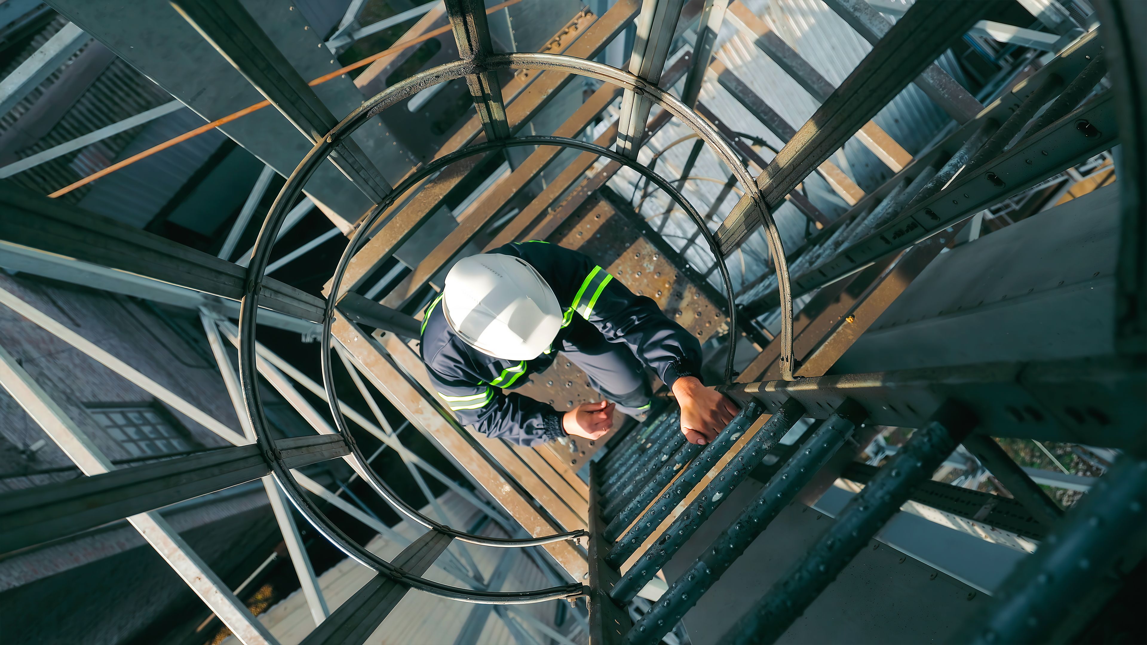 Worker Climbing Industrial Ladder with Safety Gear. Top view of a worker in a hard hat and reflective gear climbing a caged industrial ladder. Worker Climbing Industrial Ladder with Safety Gear. Top view of a worker in a hard hat and reflective gear climbing a caged industrial ladder.