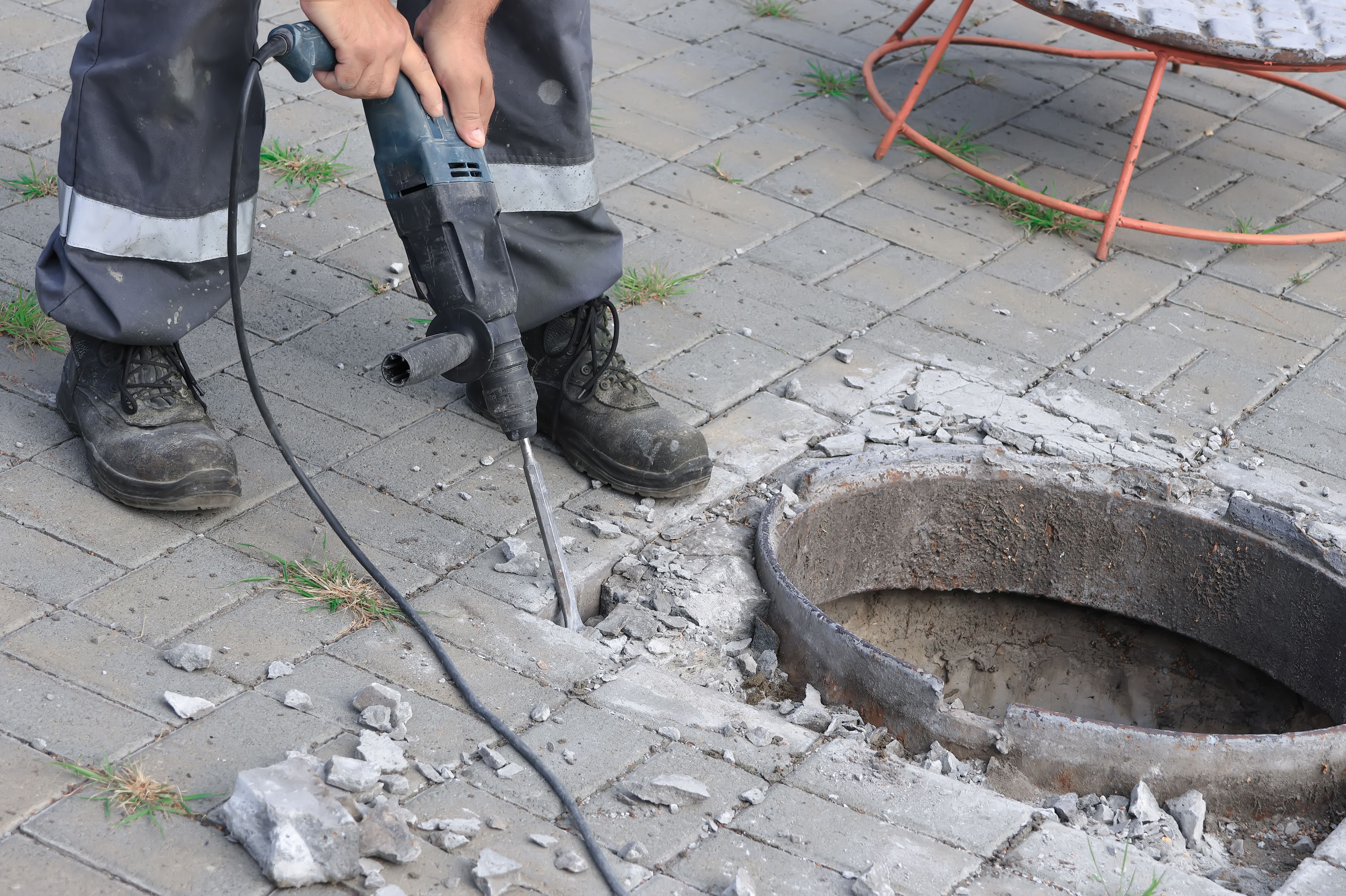 Worker using power drill on pavement near round hole, breaking concrete, protective gear