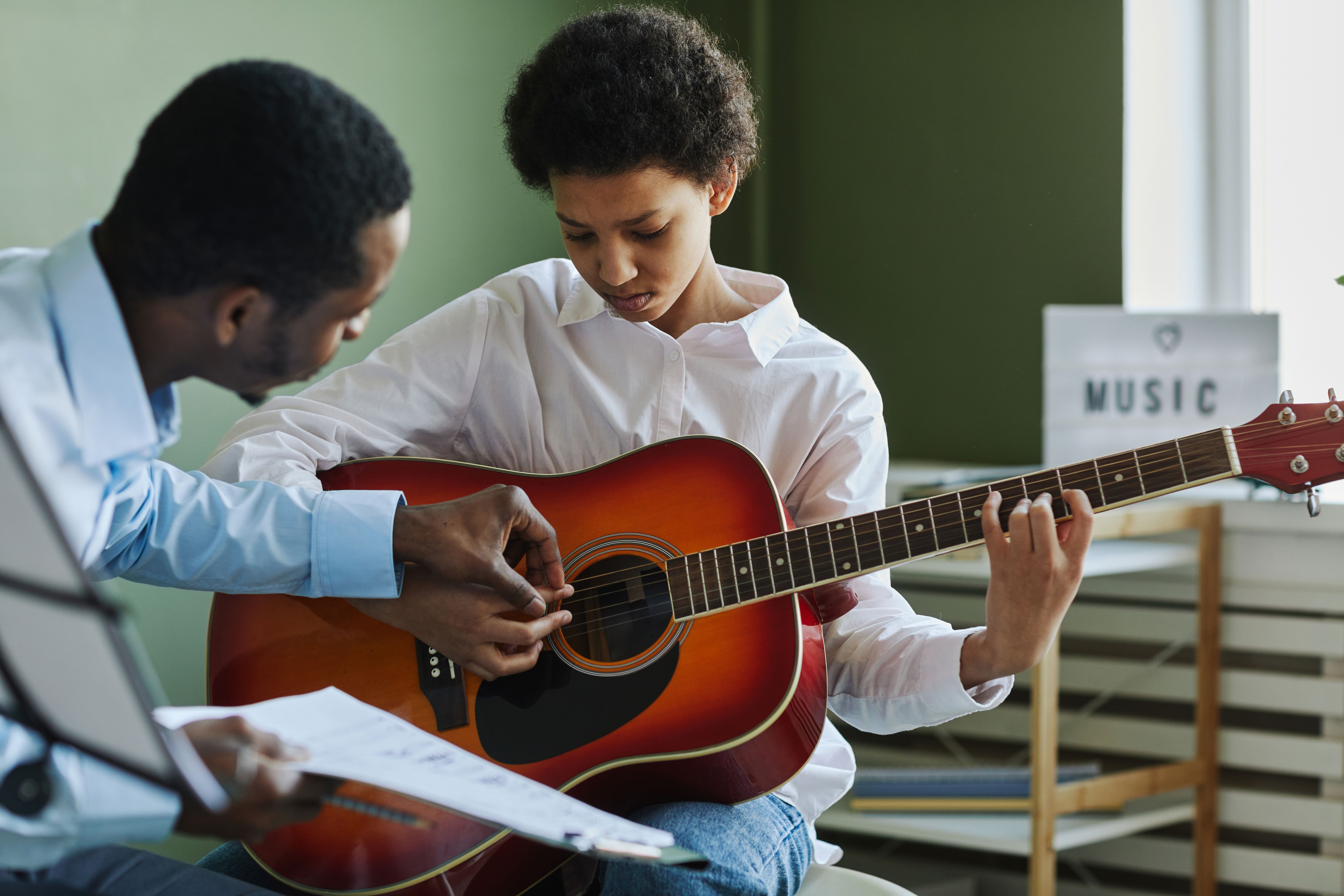 Joven y diligente que toca la guitarra acústica mientras la maestra la consulta Joven y diligente que toca la guitarra acústica mientras la maestra la consulta