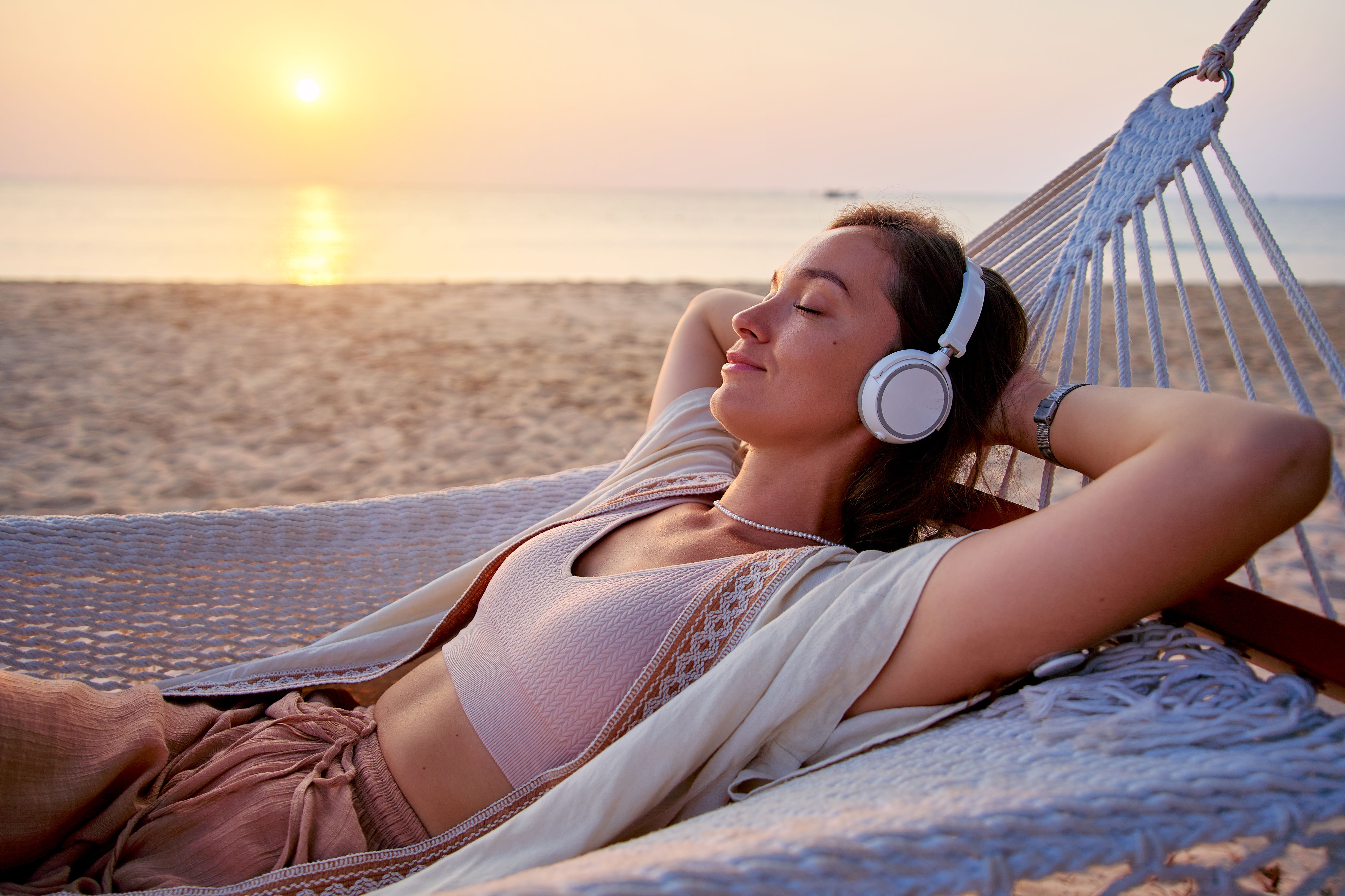 Young blissful calm woman wearing wireless headphones with closed eyes and hands behind head lie in a hammock and enjoy relaxing music Young blissful calm woman wearing wireless headphones with closed eyes and hands behind head lie in a hammock and enjoy relaxing music