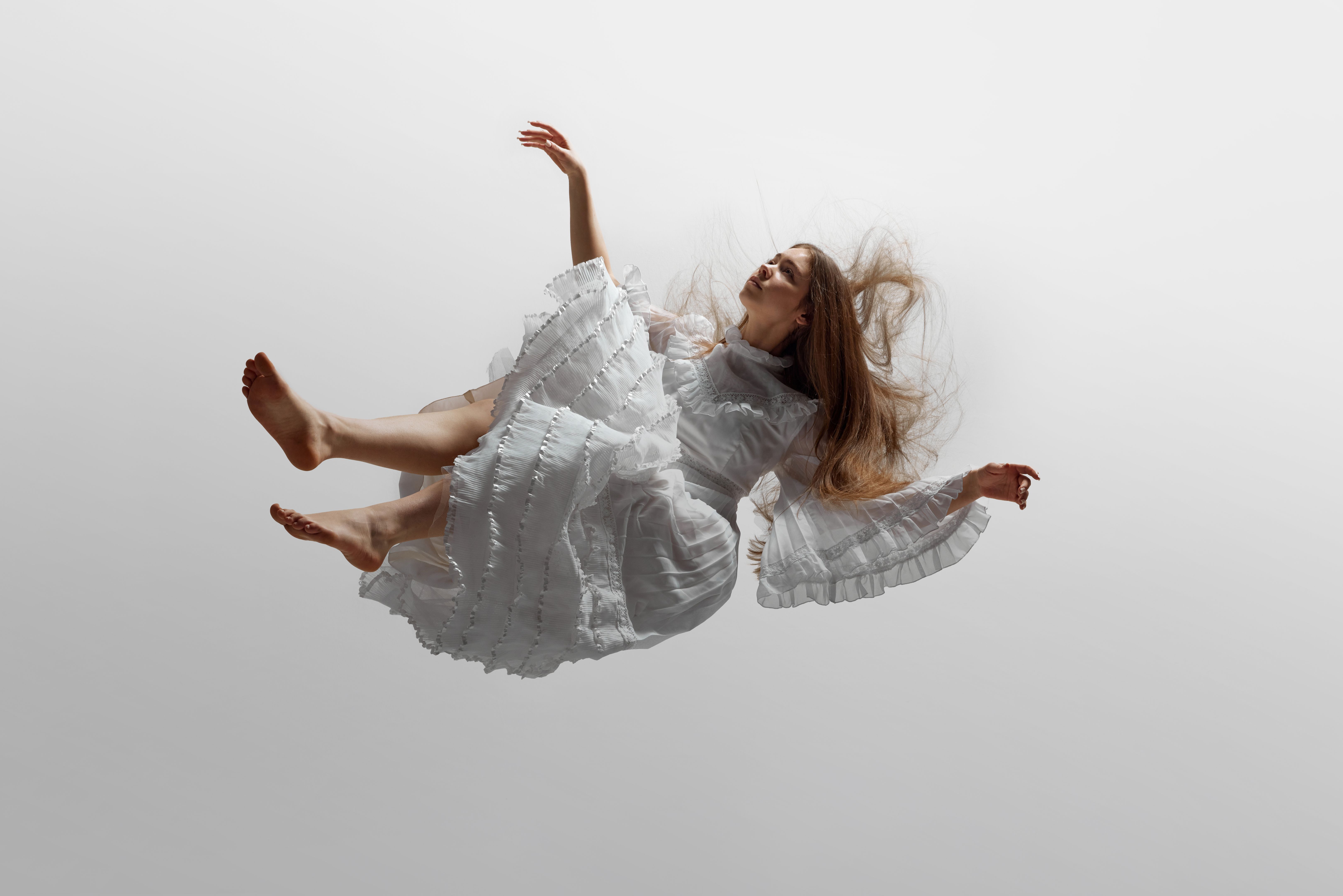 Young woman, dressed in elegant white dress with delicate ruffles, seems to levitate against white studio background.