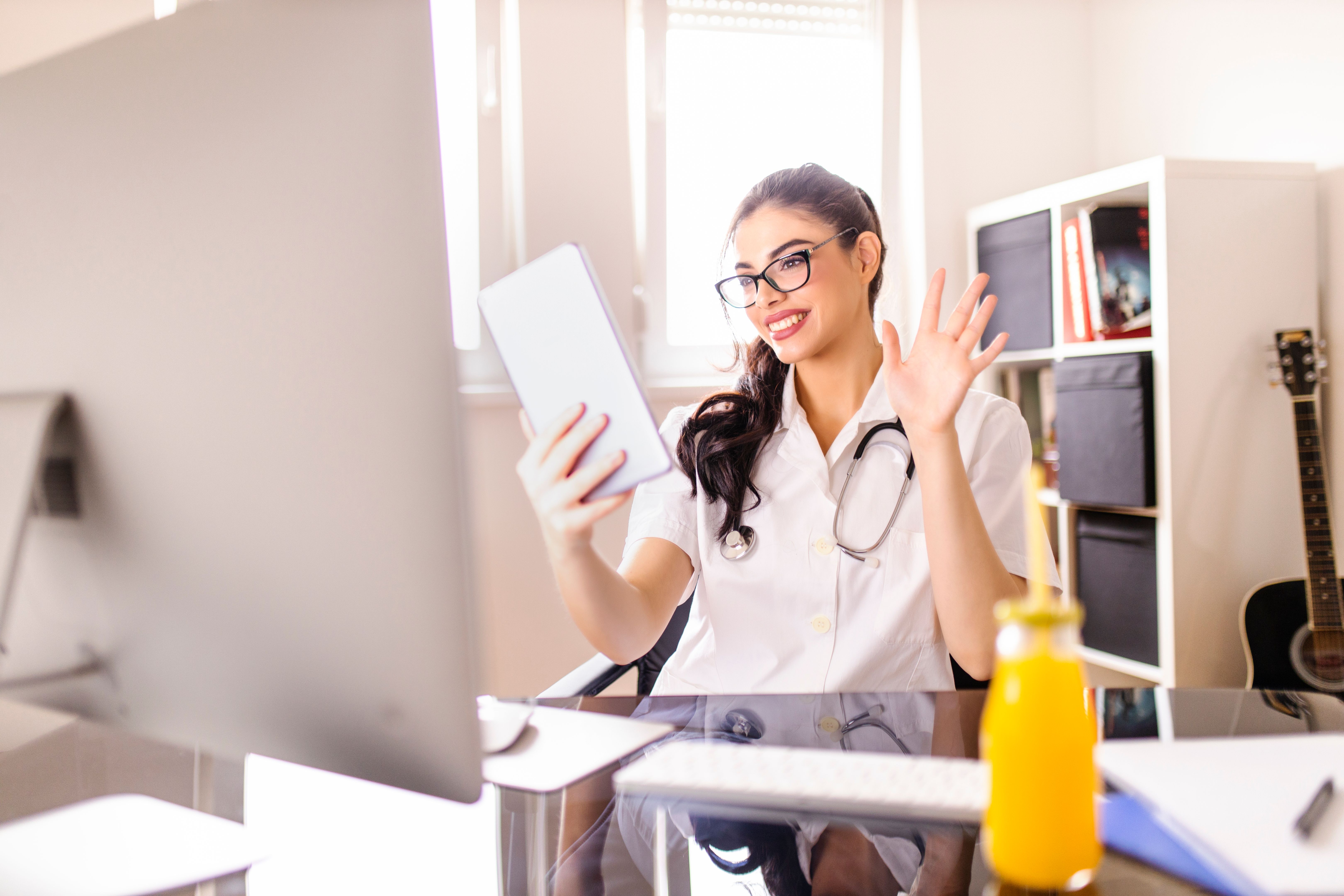 Médico trabalhando, tendo chamada de vídeo em tablet digital e bebendo suco de laranja no consultório