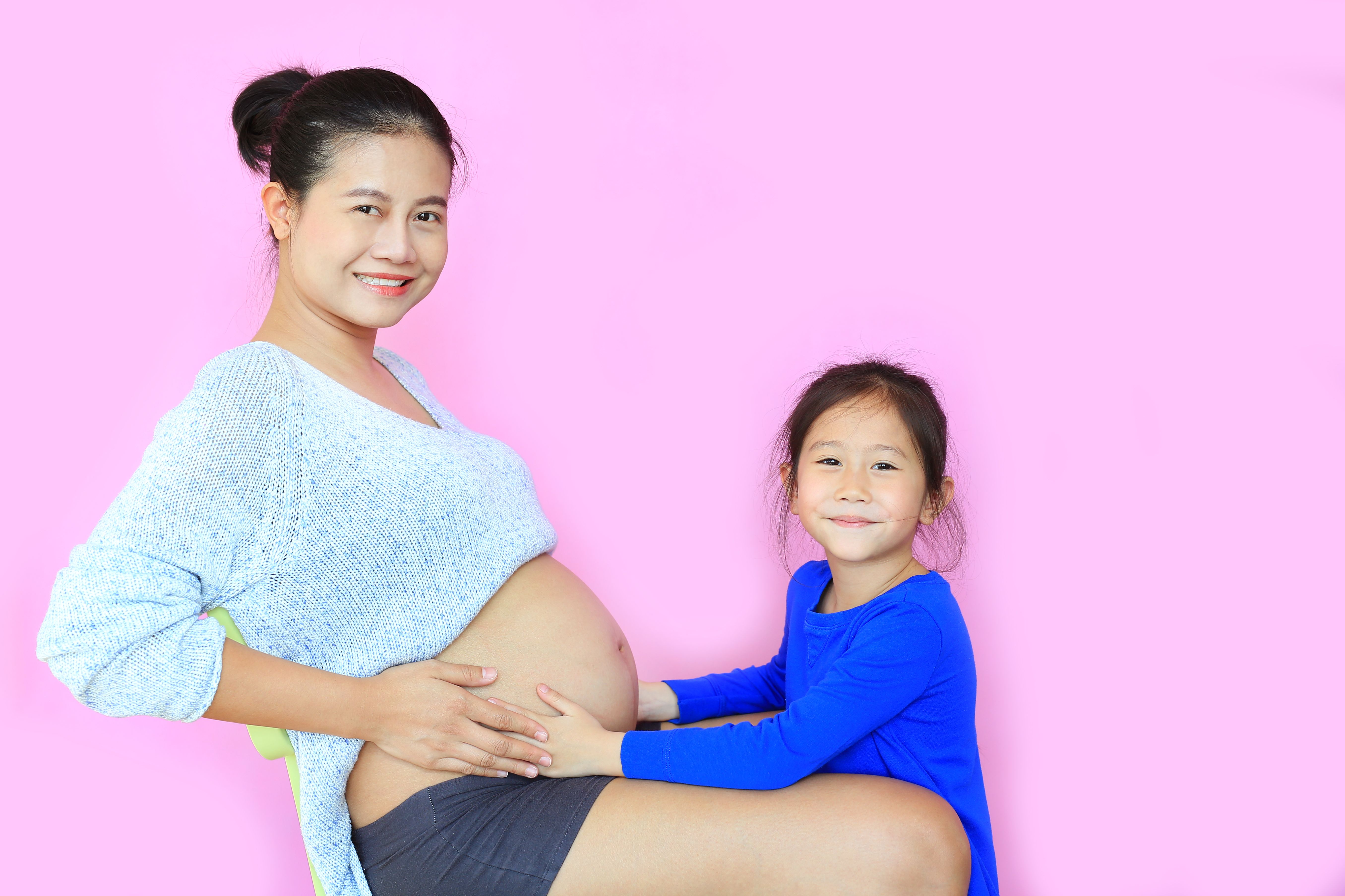 Happy asian mom and daughter with love. Little child girl relaxing with pregnant mother isolated on pink background. Pregnancy and new life concept.