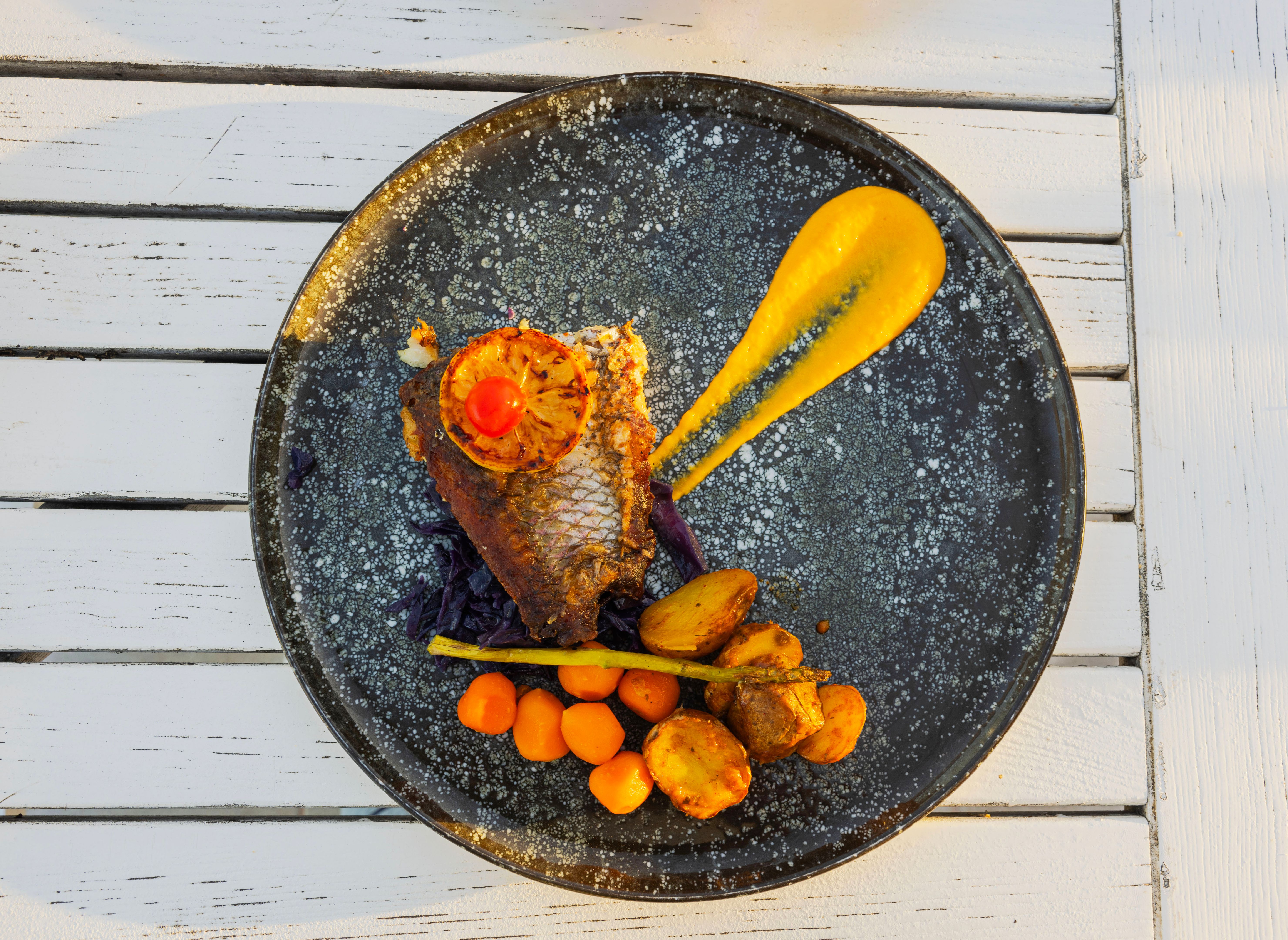 Grilled fish fillet with lemon, cherry tomato, carrots, potatoes and yellow sauce served on black ceramic plate. Curacao.