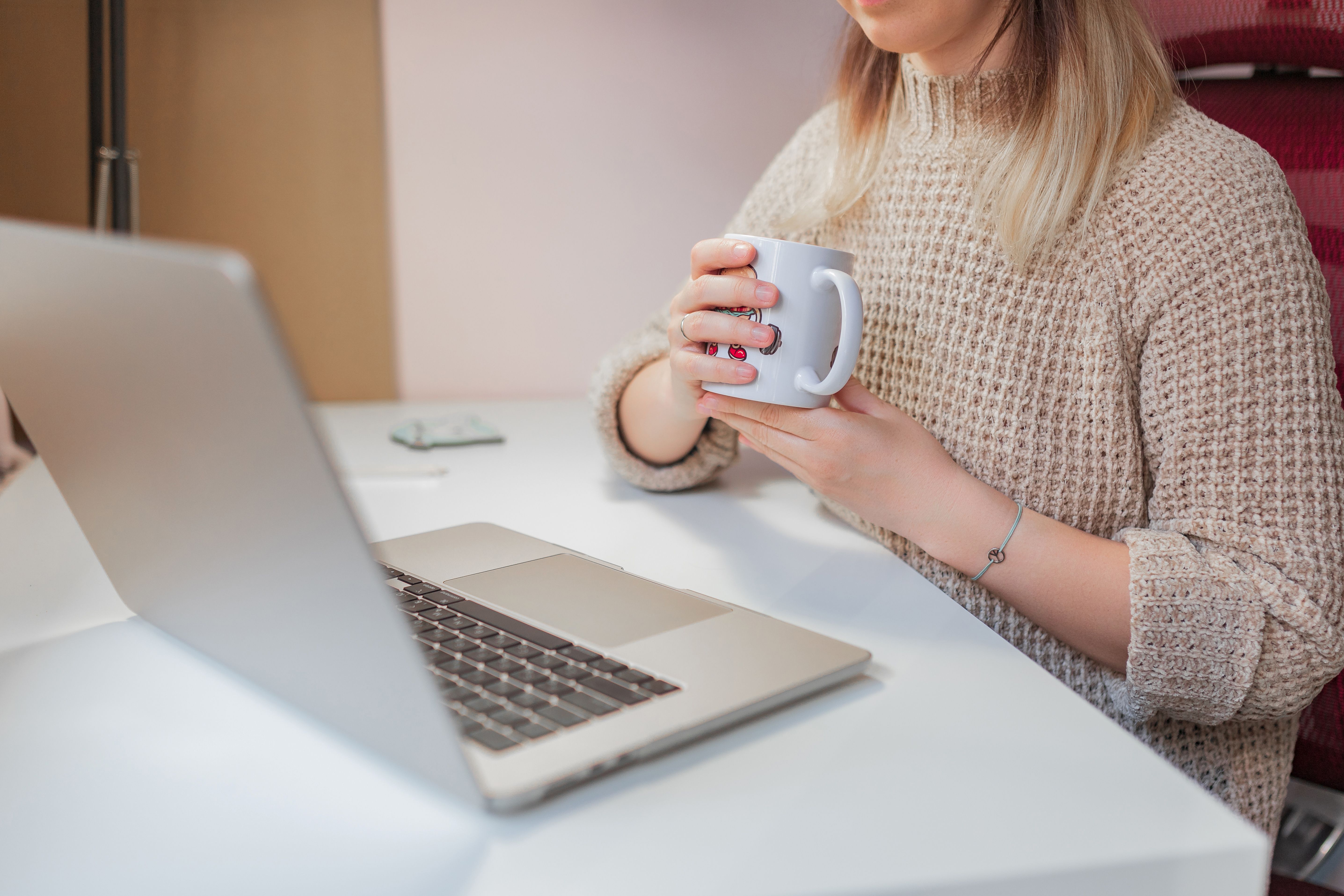 Woman holds mug while seated at desk with open laptop during work break.