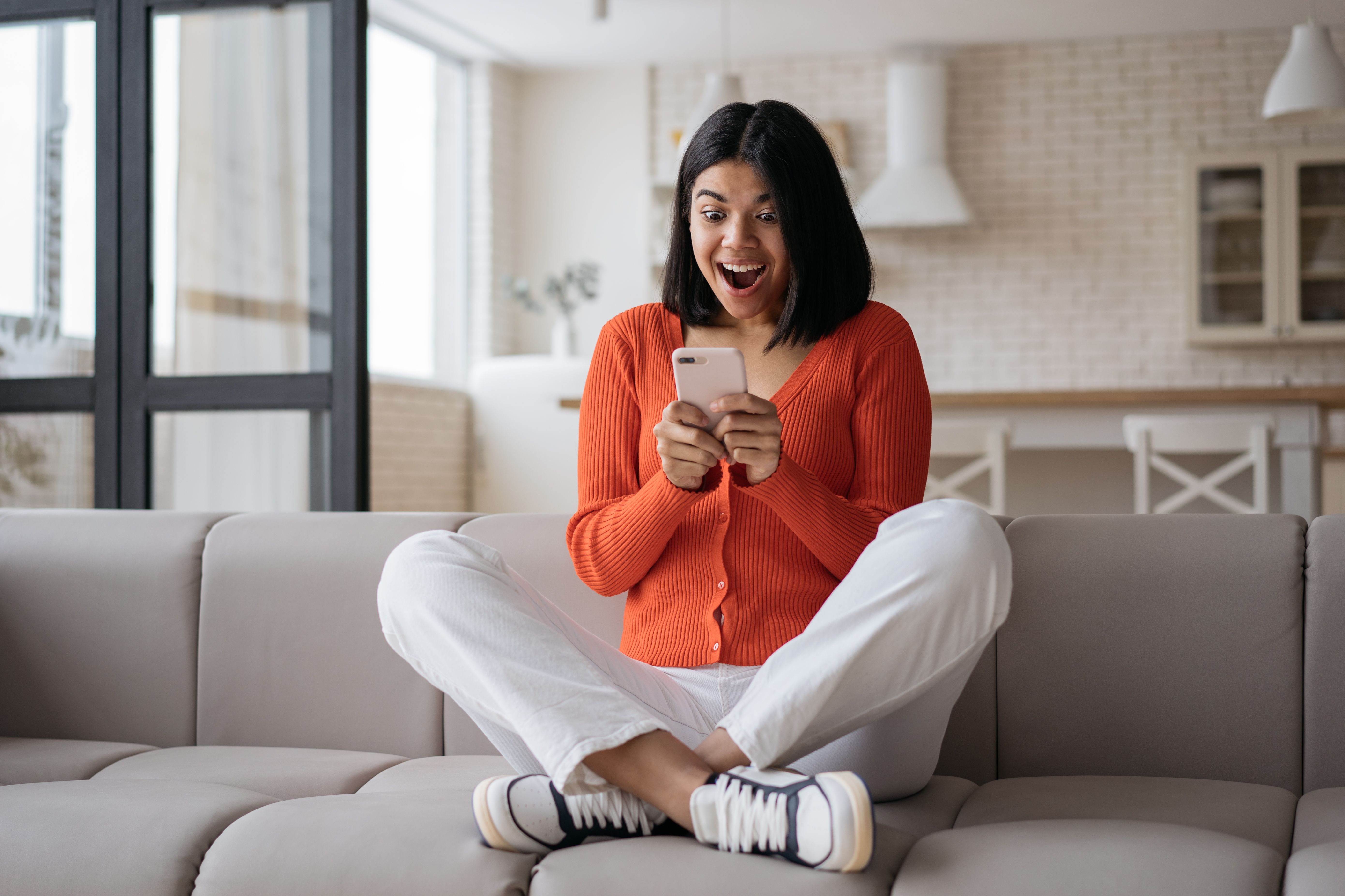 Woman holding smartphone playing mobile game celebration success at home. Excited African American hipster using mobile phone shopping online with big sales sitting on sofa