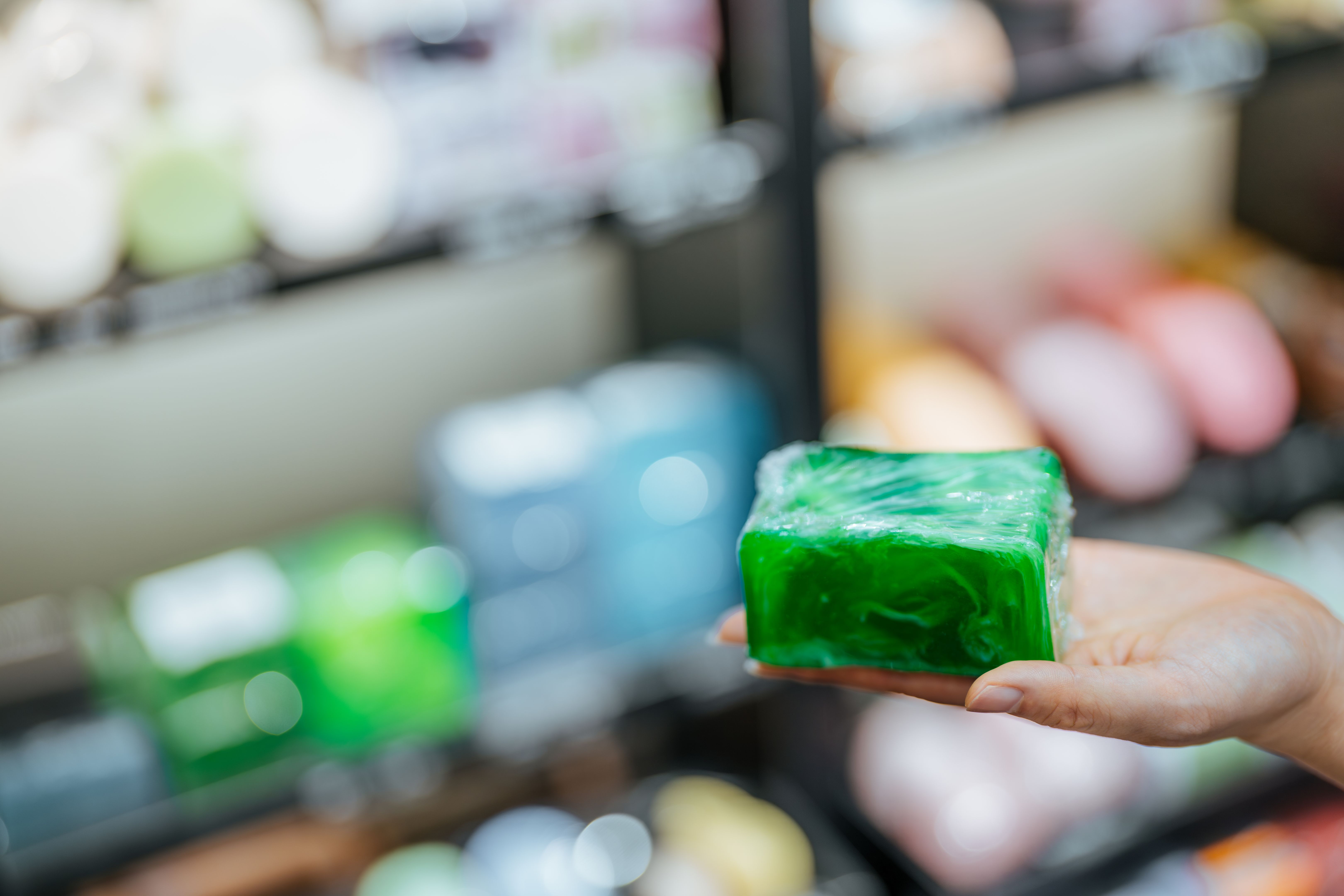 Close up of Asian young woman shopping for handmade soap.  Hand with lavender herbal soap on the background of shelves with a range of products for body care and bathroom cosmetics