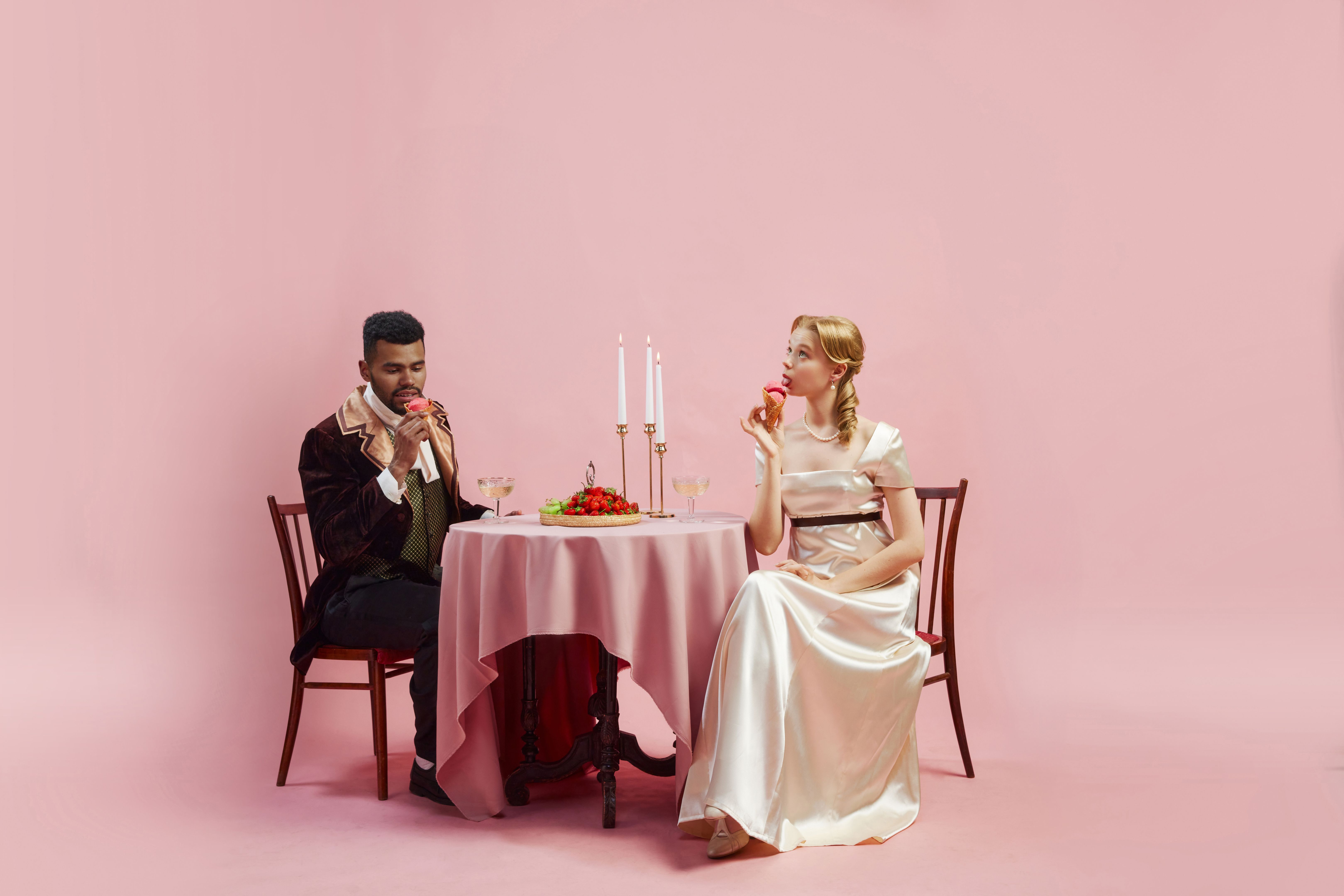 Handsome young man and elegant young woman in period costume spotting at table and eating ice cream against pink studio background
