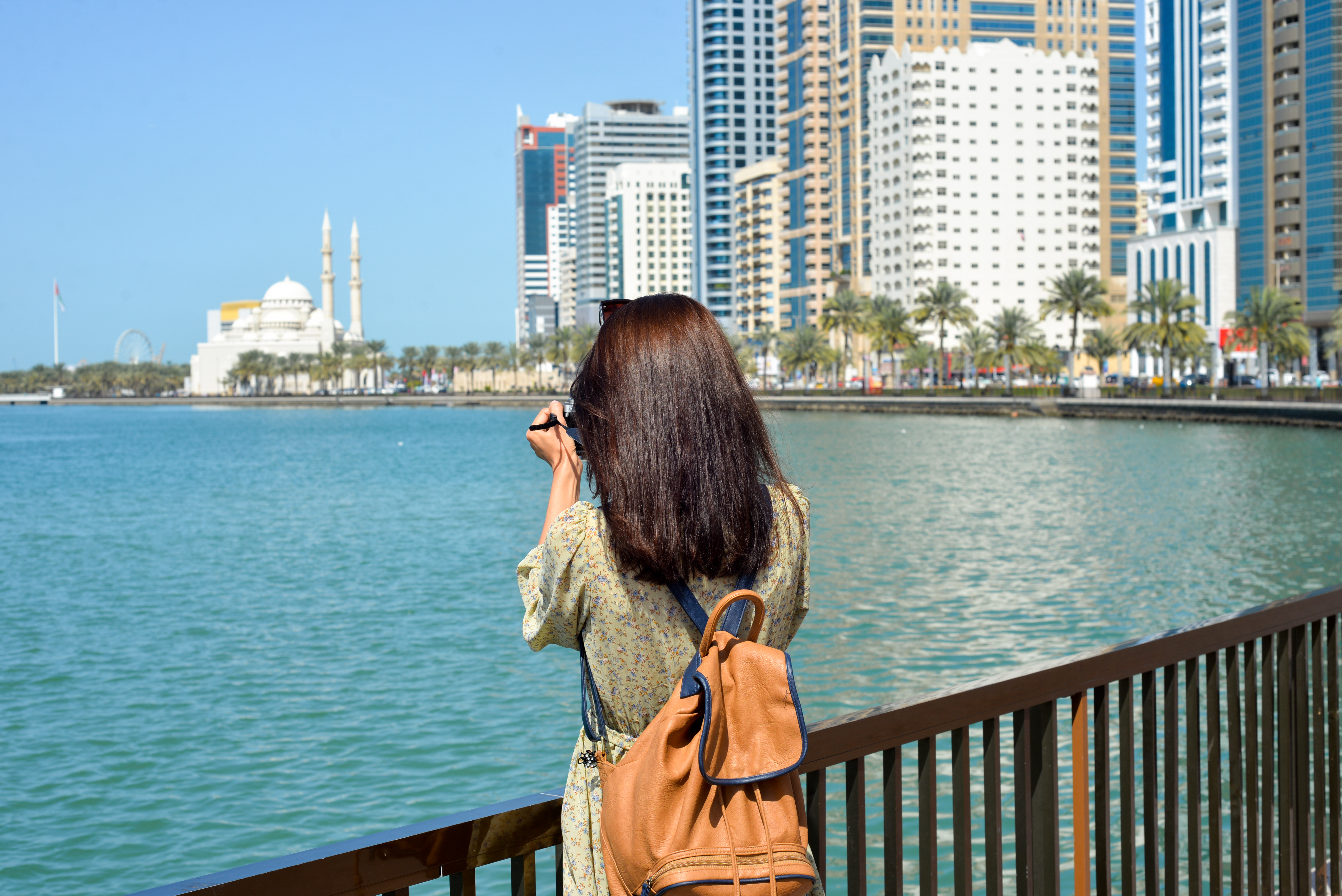 A woman in a long dress with a backpack walks with a camera along the Al Majaz embankment, Lake Khaled, Sharjah emirate. Rear view of a woman photographing the embankment