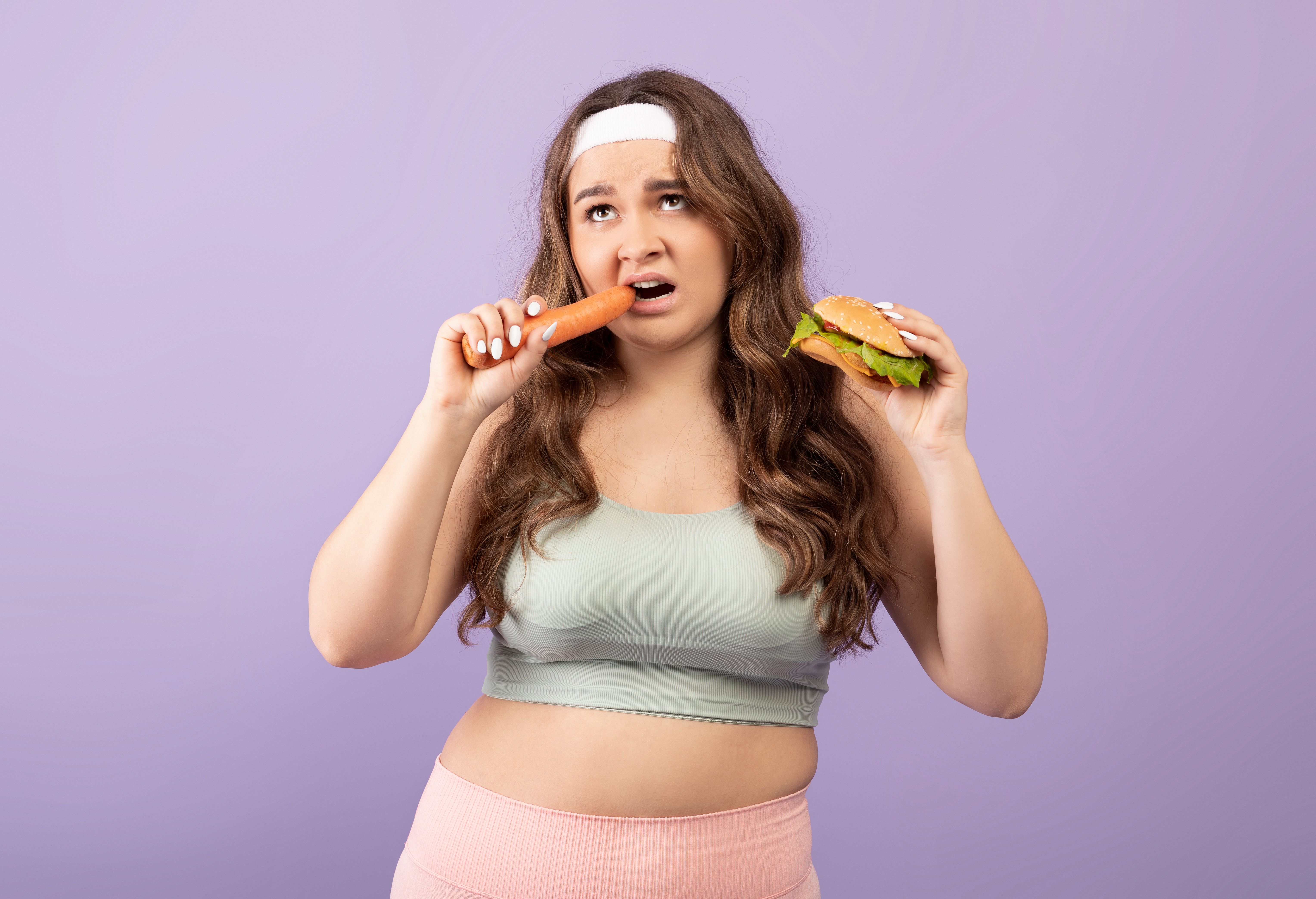 Depressed plus size european millennial female in sportswear eats carrot and holds burger
