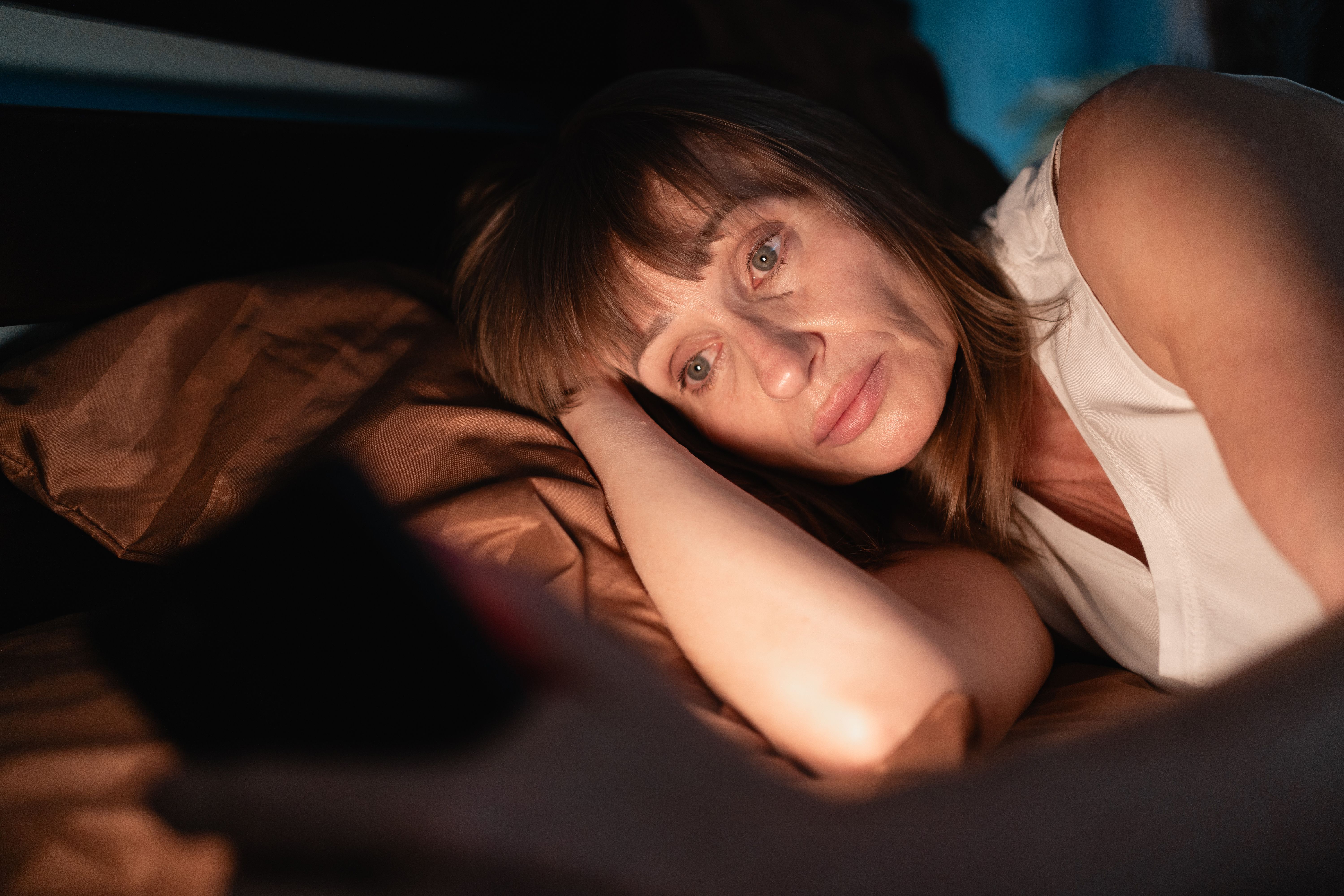 Sleepy woman awake at night using smart phone lying in bed in her bedroom. Using mobile for chatting