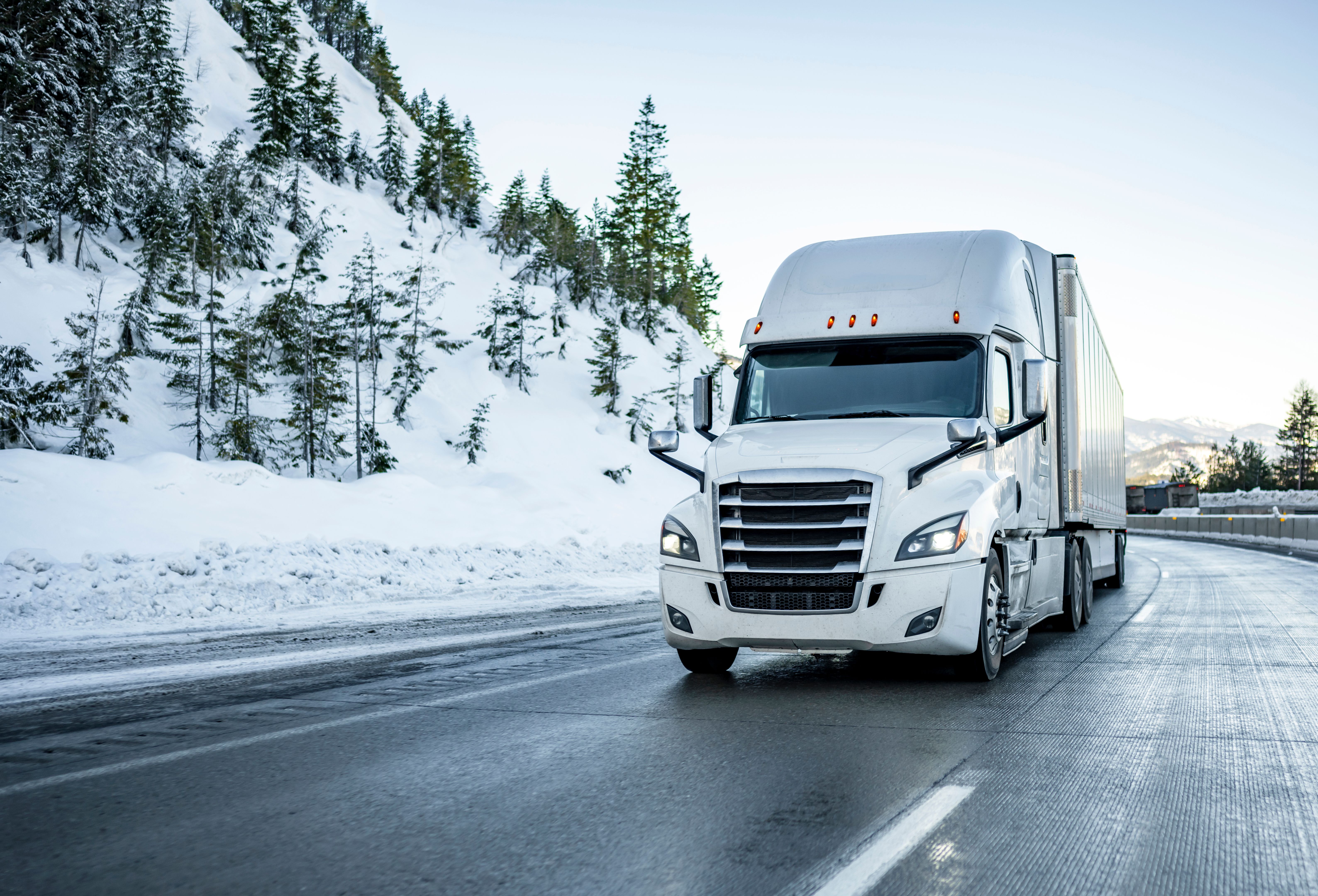 Popular white big rig semi truck tractor transporting cargo in dry van semi trailer running on the iced winter highway road with covered by snow hill of the mountain in Montana