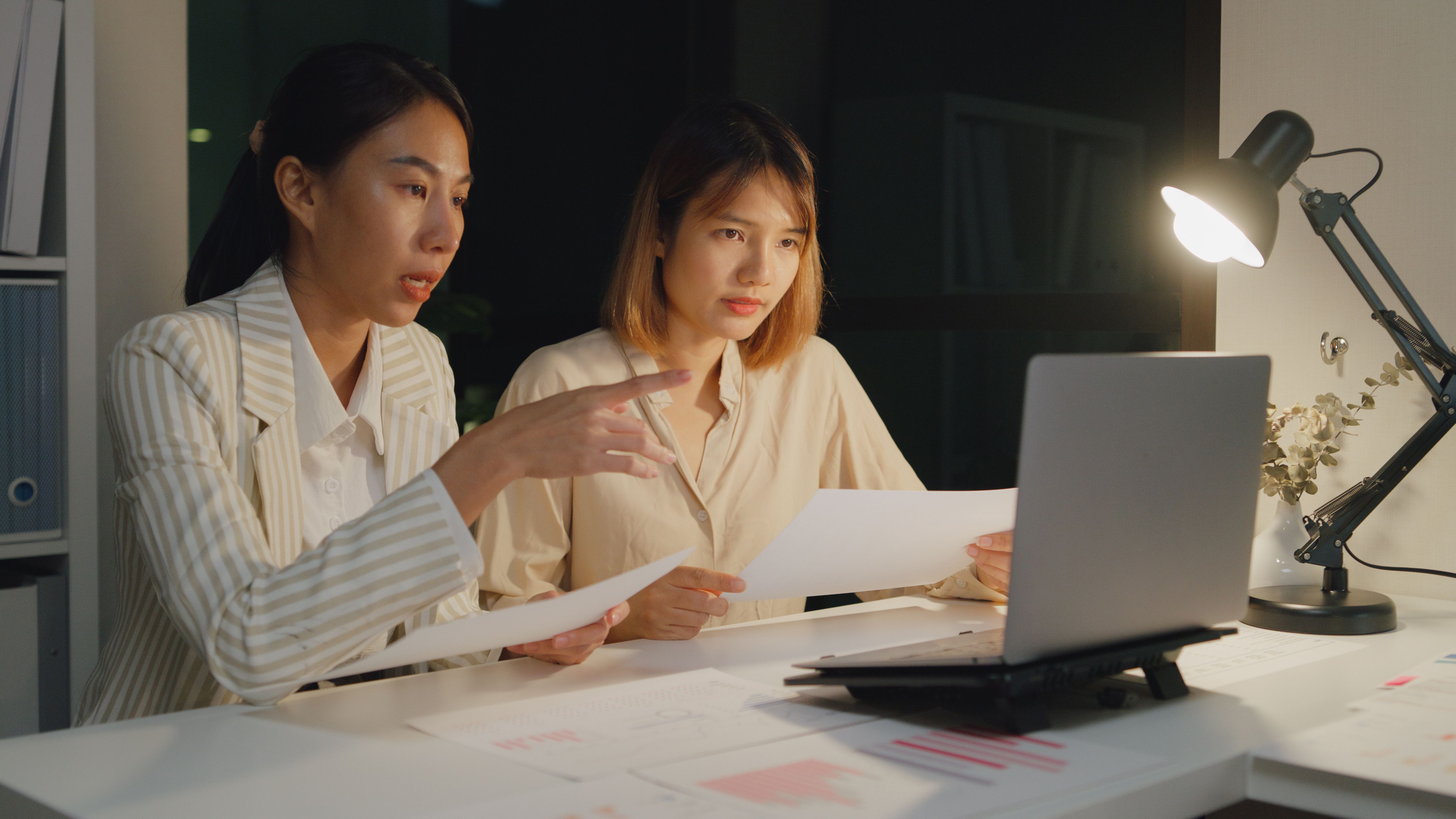 Young Asian businesswomen sit in front of computer laptop look at screen discussing chart reporting in presentation and compare data overtime working at night. Working late. Young Asian businesswomen sit in front of computer laptop look at screen discussing chart reporting in presentation and compare data overtime working at night. Working late.
