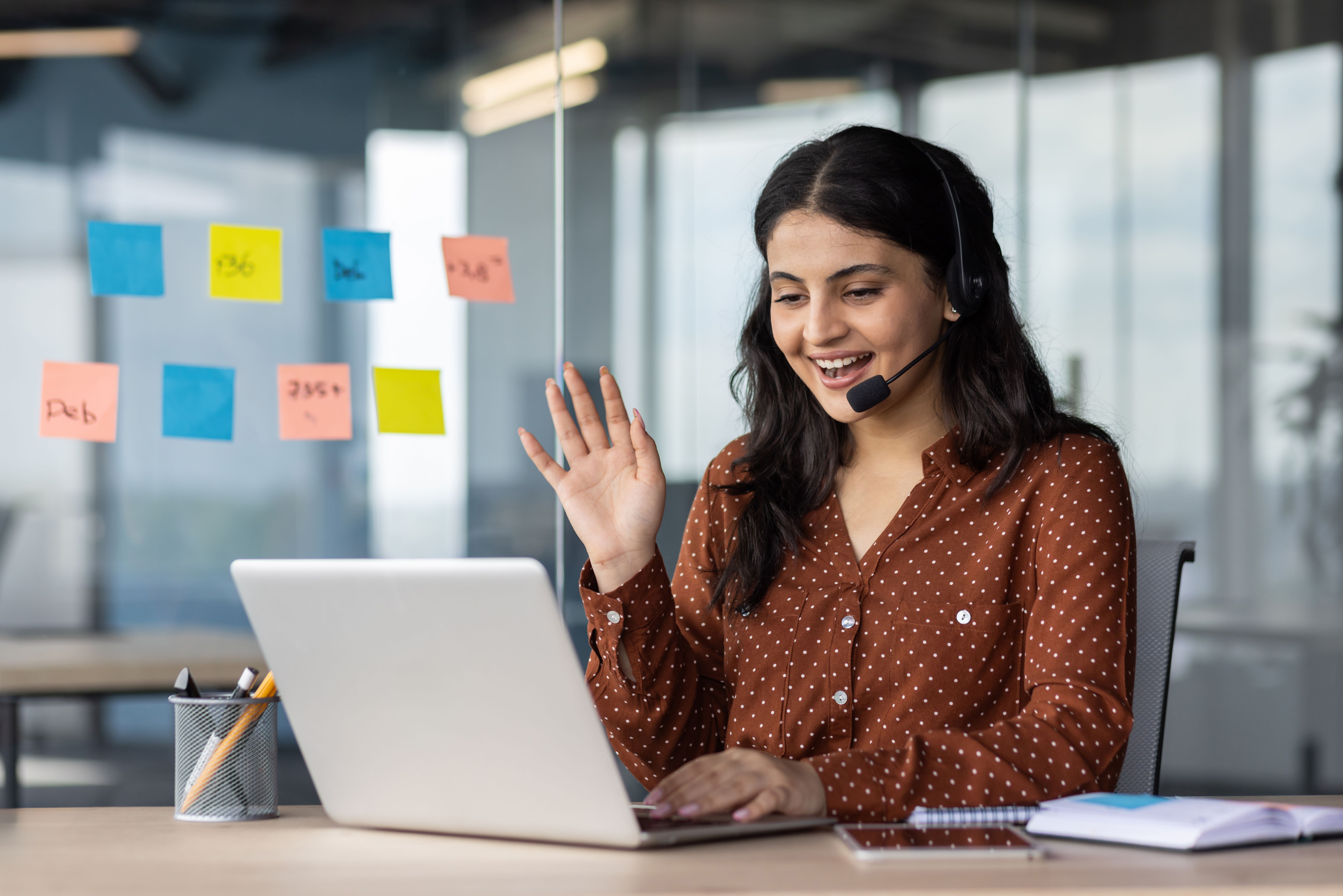 Latin American woman with headset phone using laptop for video call, female online customer support worker waving hand greeting customer, online meeting with colleagues remotely