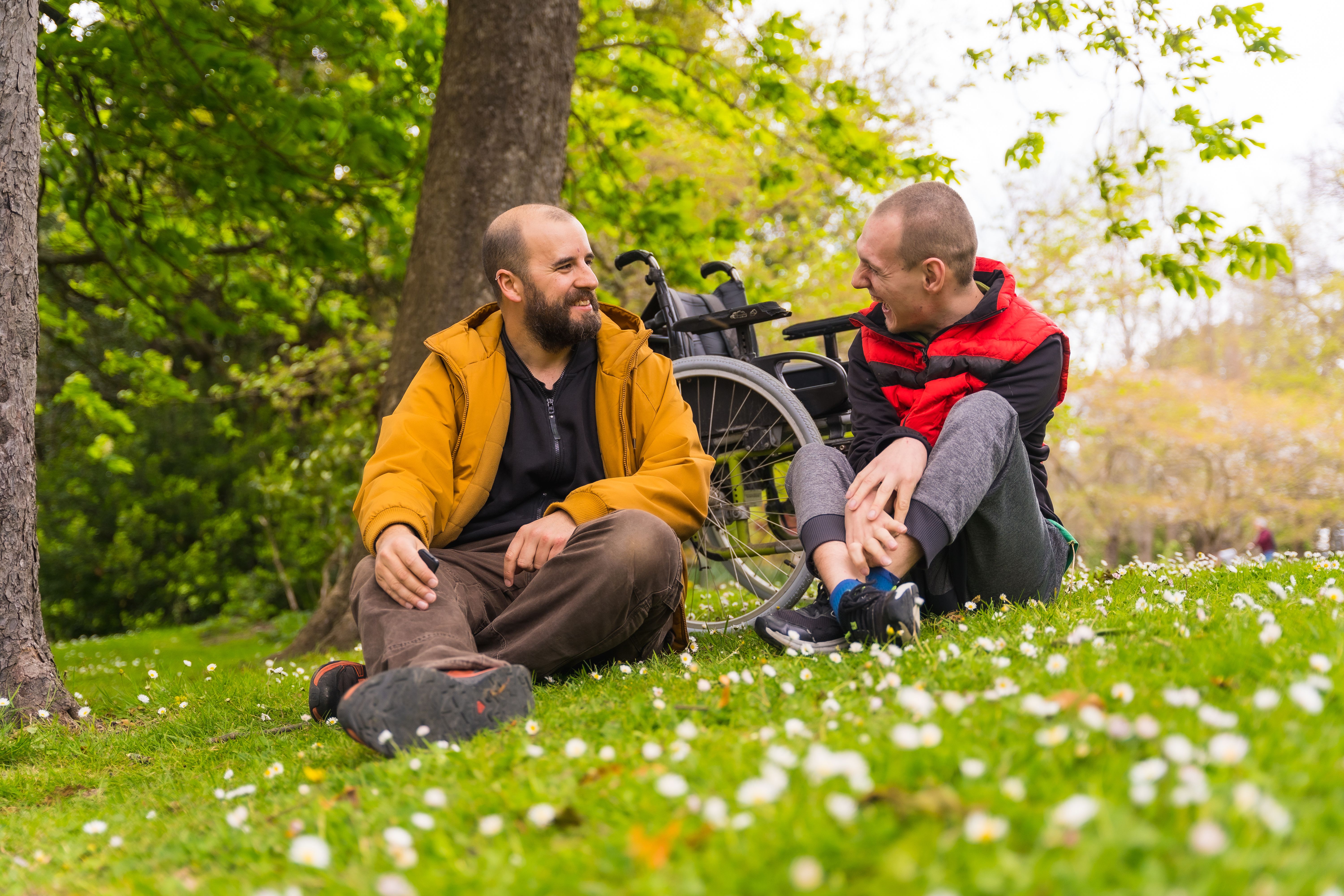 A paralyzed young man with a friend sitting on the grass of a public park in the city, talking and laughing