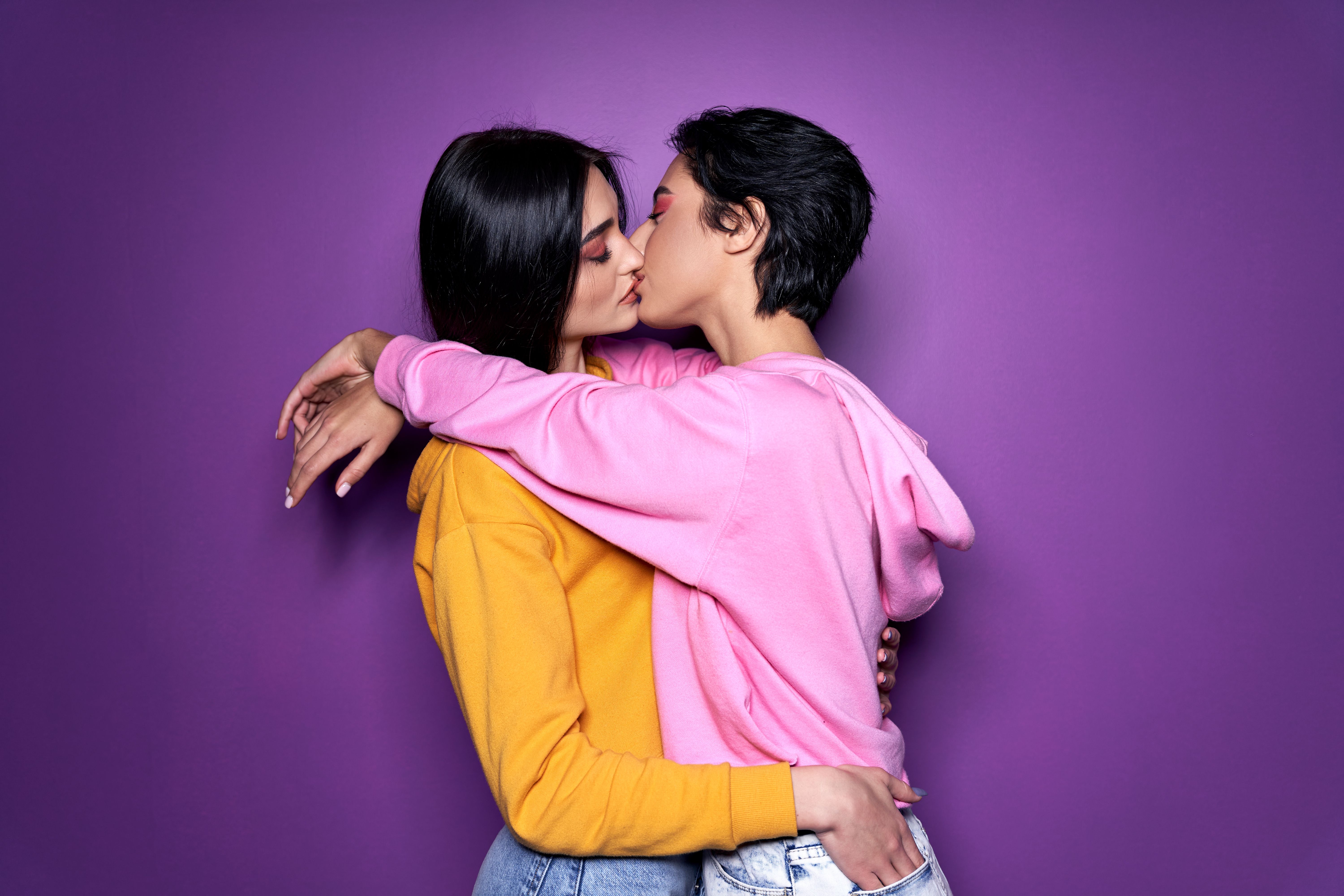 Two pretty sexy stylish cool generation z girls lgbtq lesbian couple dating in love hugging enjoying intimate tender sensual moment together kissing with eyes closed isolated on purple background.
