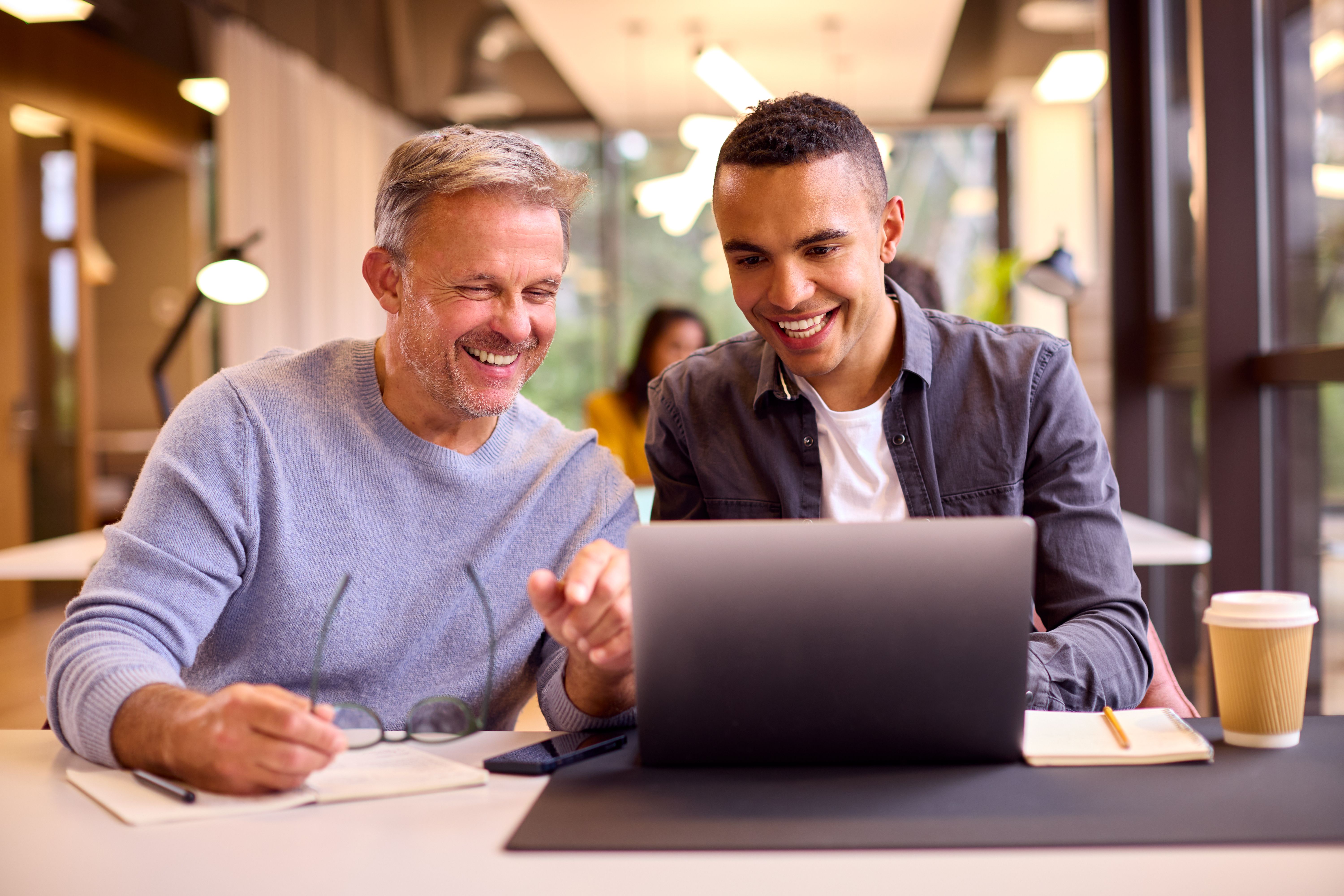 Mature Businessman Mentoring Younger Colleague Working On Laptop At Desk