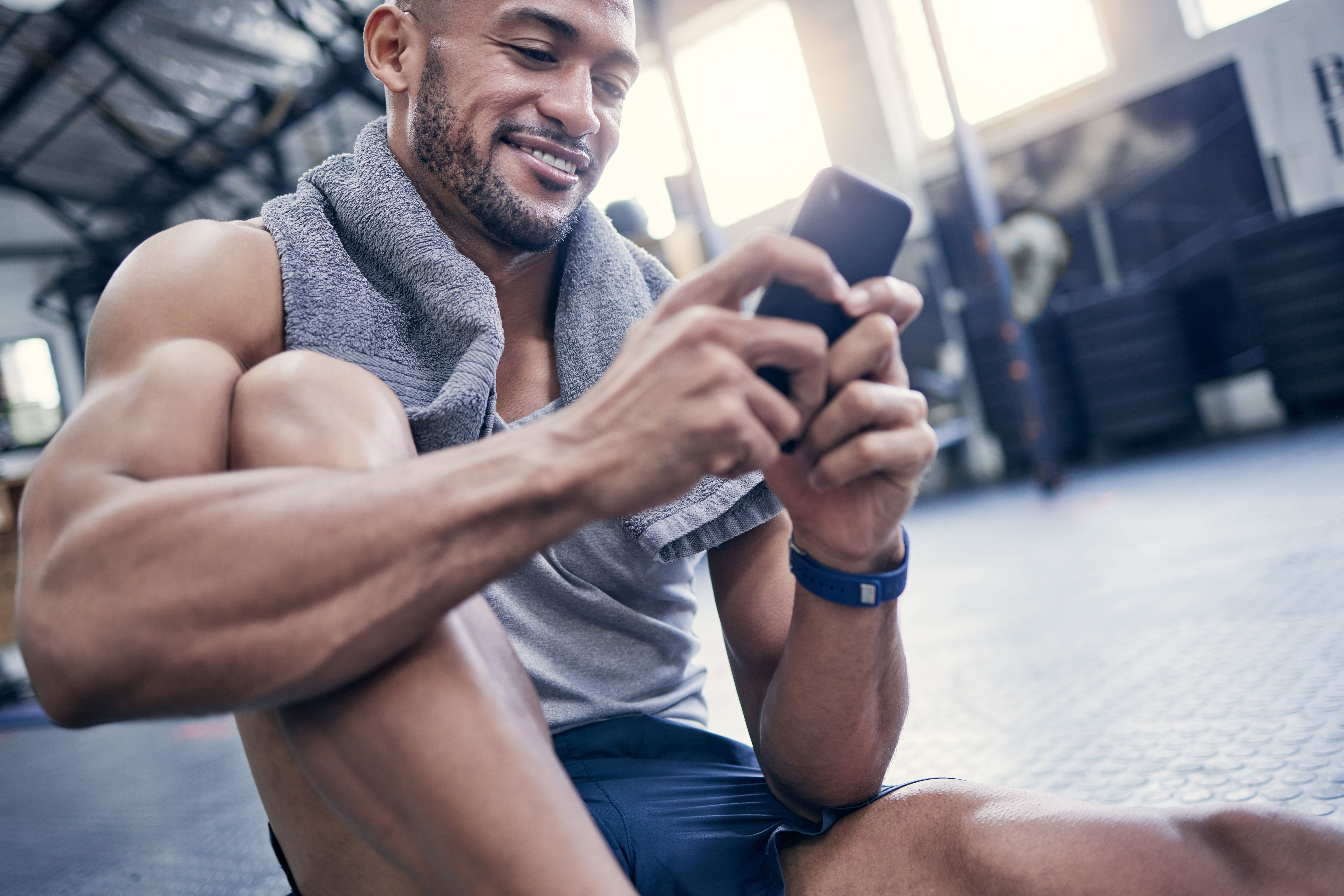 Shot of a muscular young man using a cellphone while taking a break from exercising in a gym