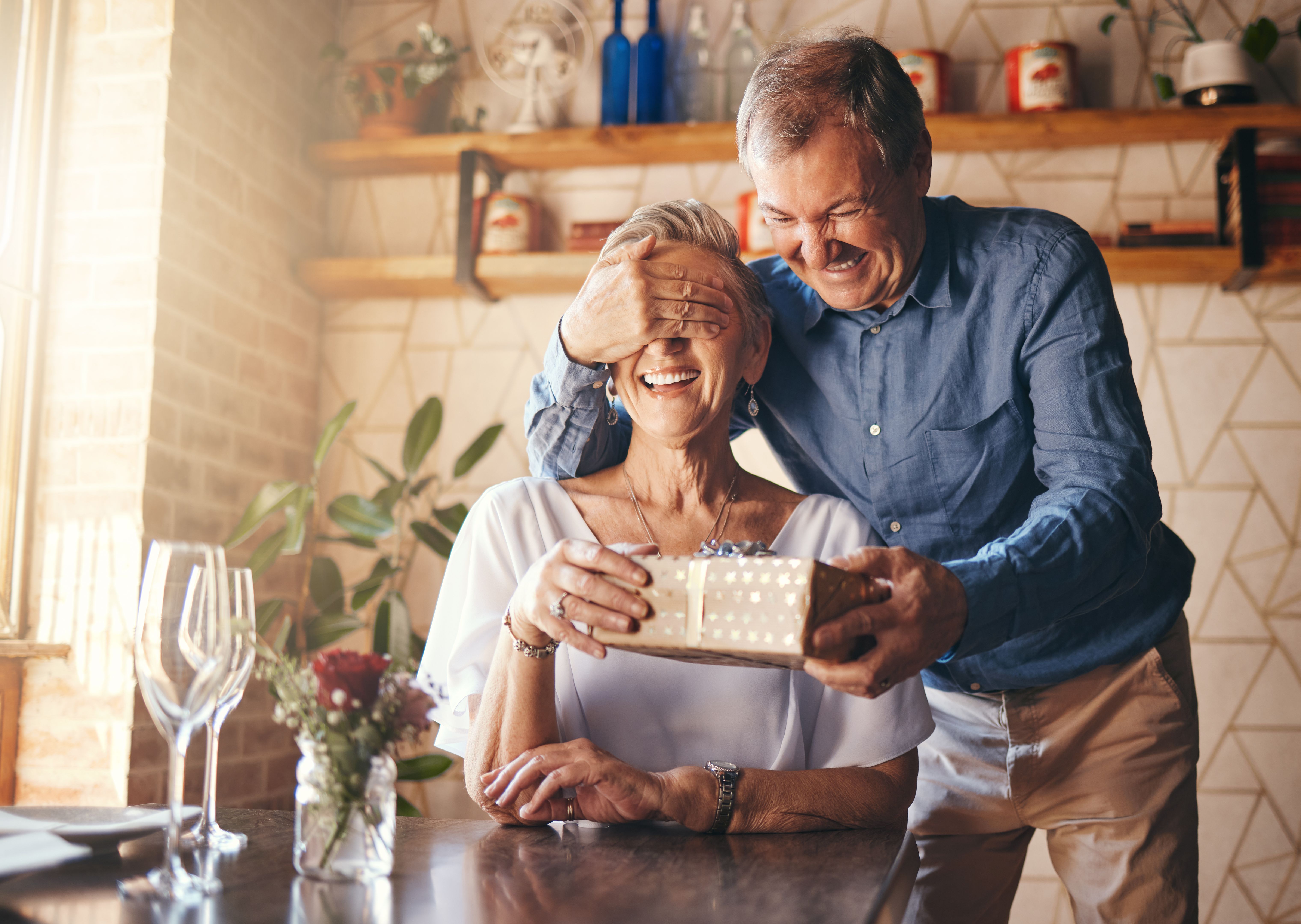 Gift, surprise and senior couple in restaurant for retirement celebration or happy birthday with wine glass, present and roses. Elderly woman with marriage anniversary event with vintage fine dining