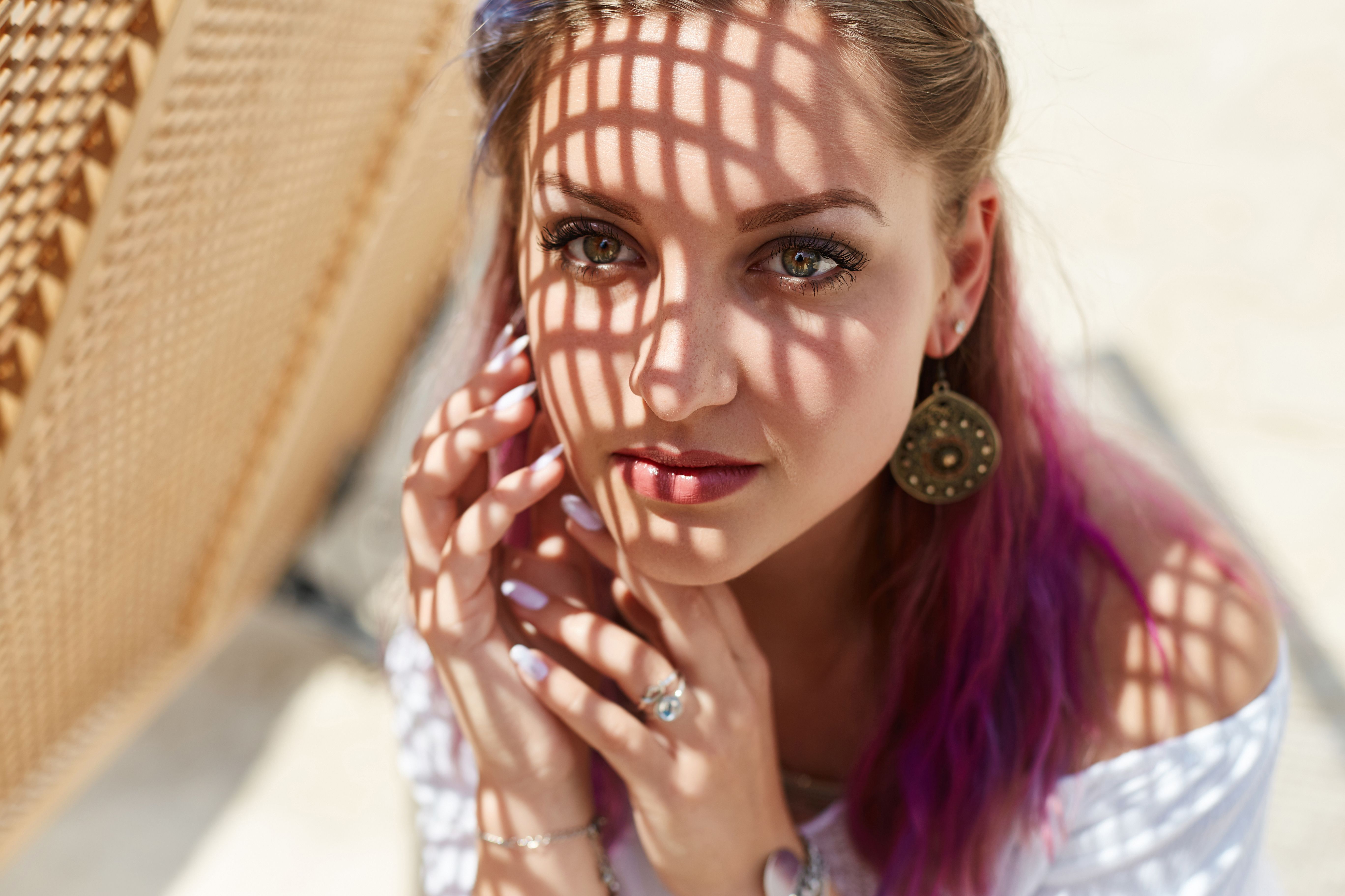 young woman with purple-tinted hair poses in an outdoor area, her face partially illuminated by intricate shadows. Bright sunlight enhances her striking features and earrings