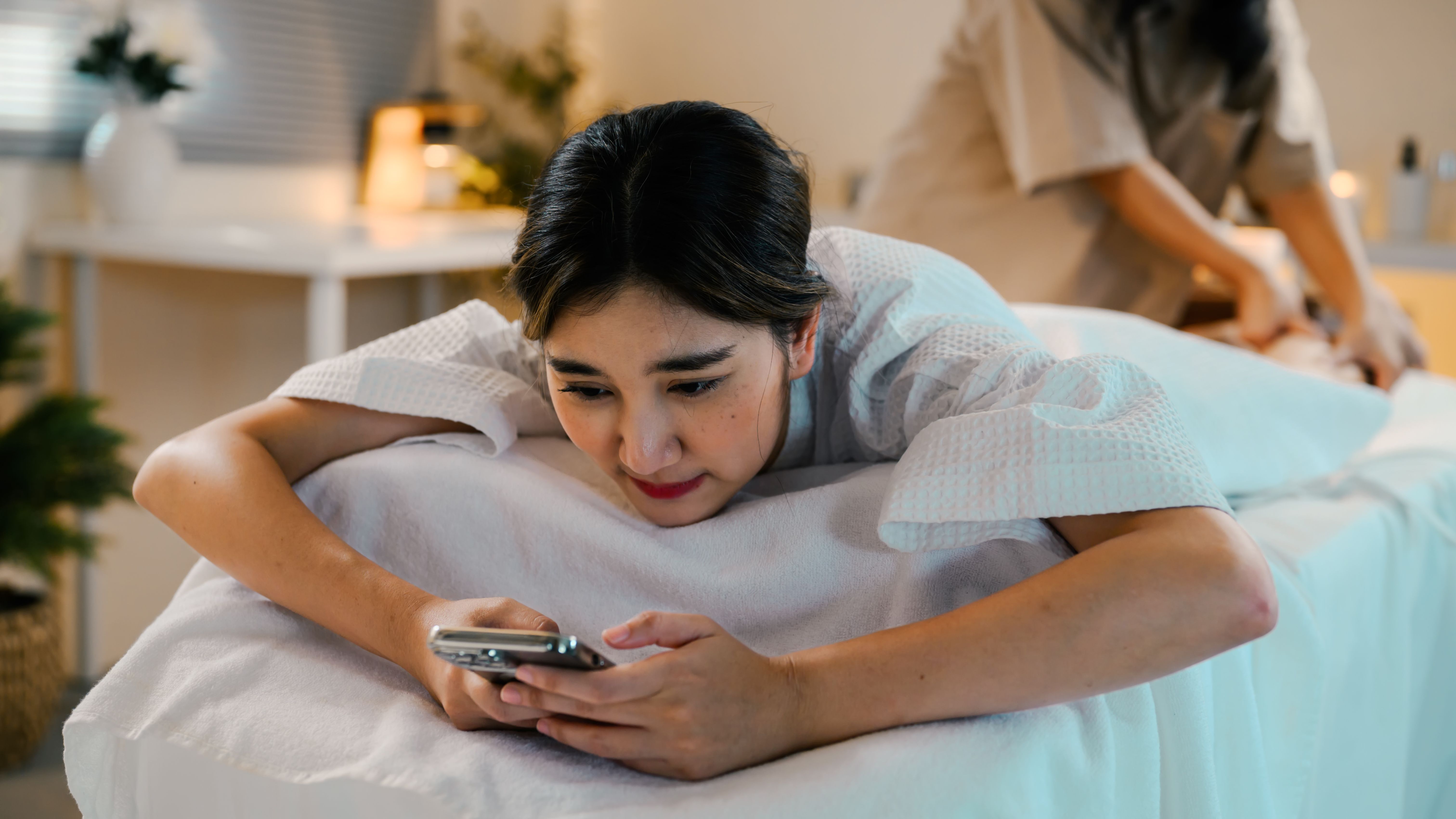 Young woman using smartphone while receiving a back massage at a spa