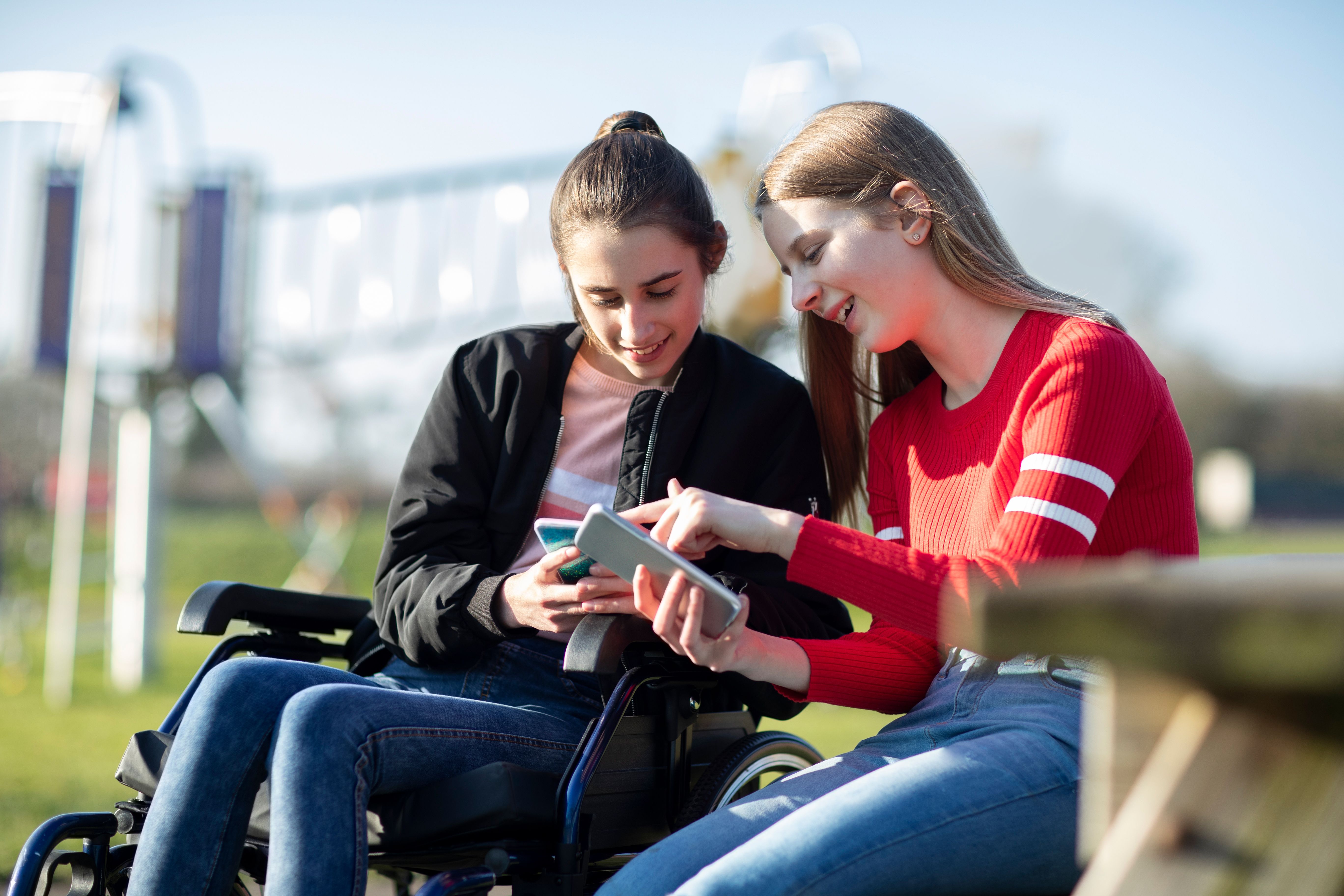 Teenage Girl In Wheelchair Looking At  Mobile Phone With Friend In Park