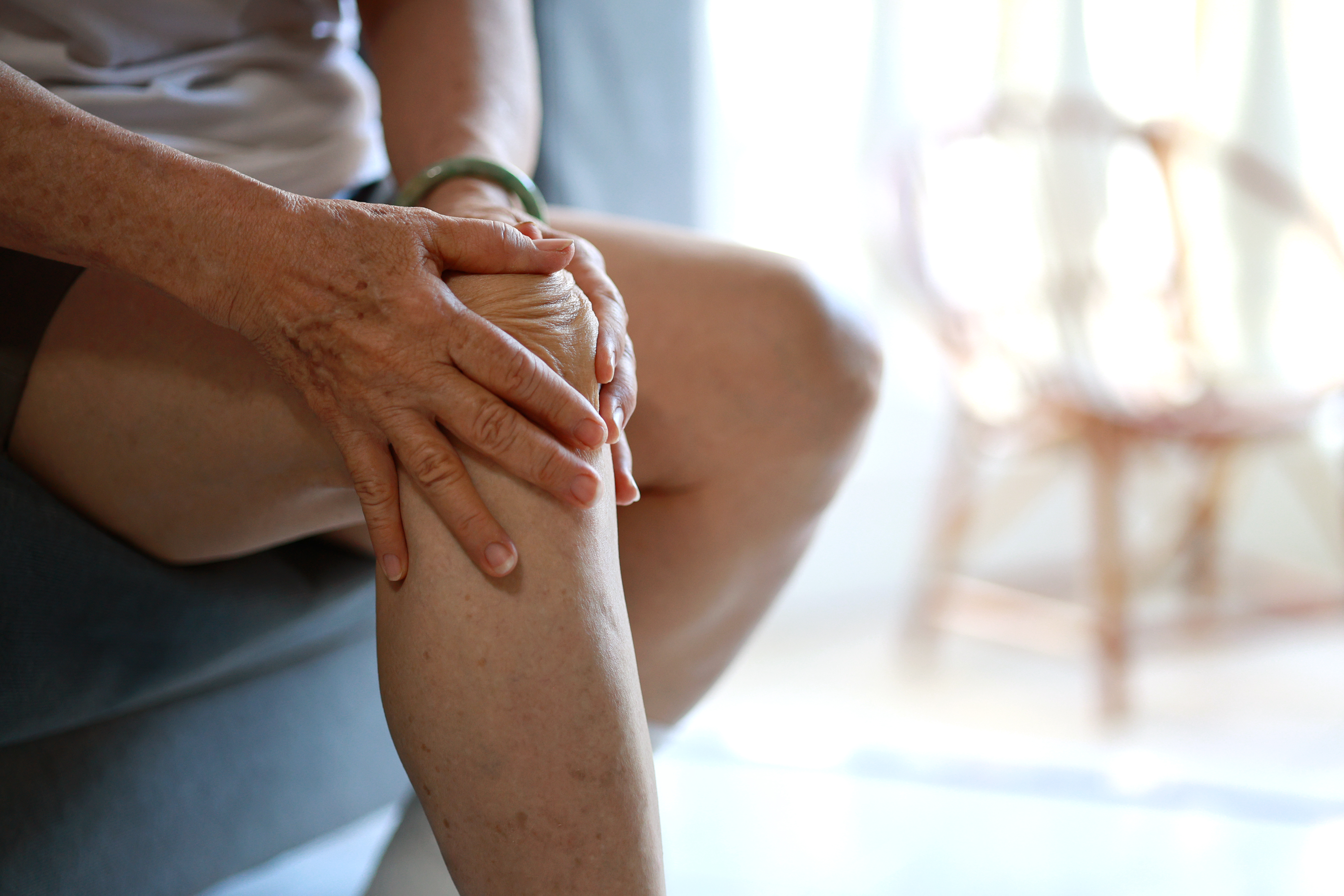 A senior Asian woman is massaging her knee while sitting on a sofa in the living room at home, visibly suffering from knee pain