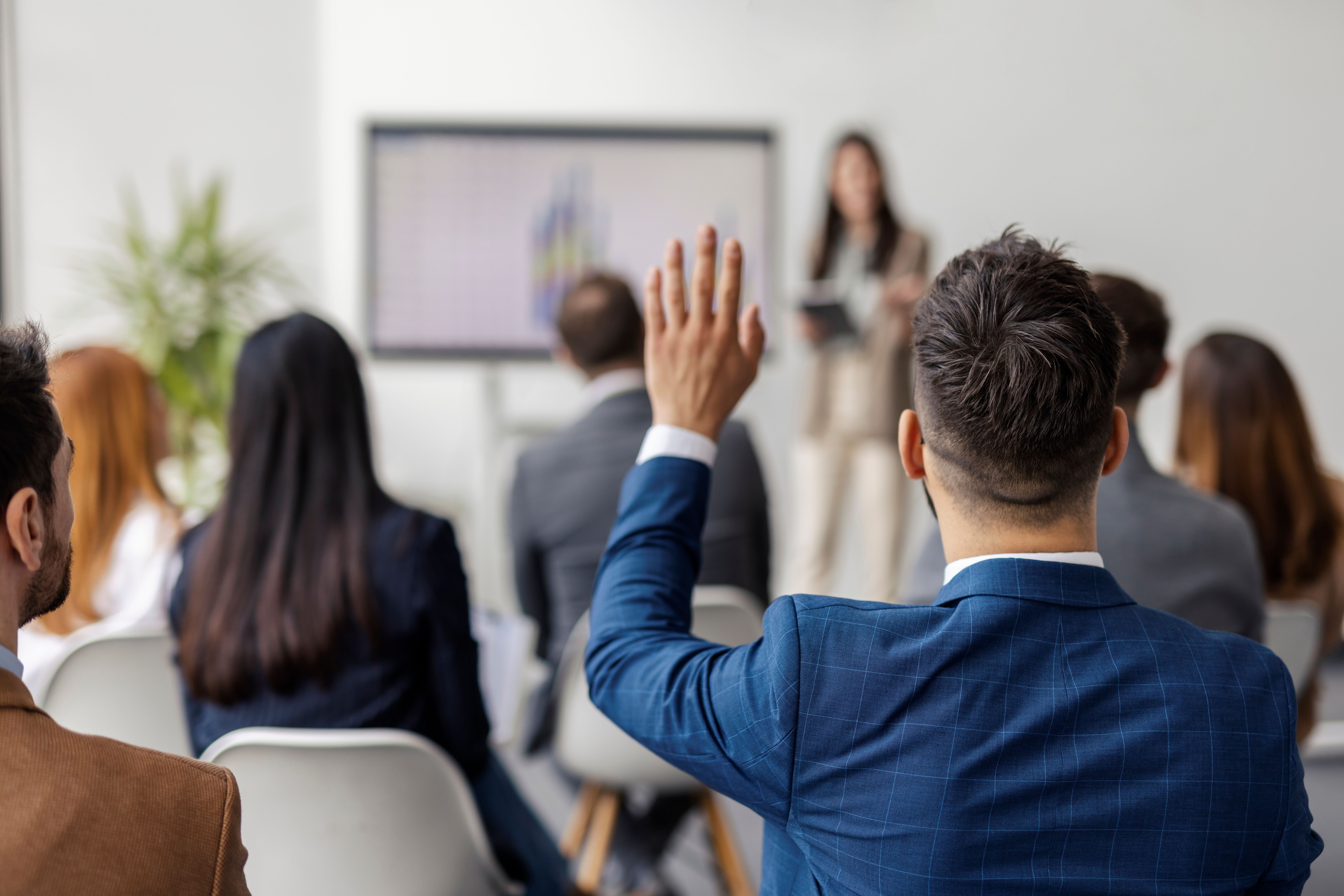 Rear view of businessman sitting in conference room on presentation with hand raised and asking a question.