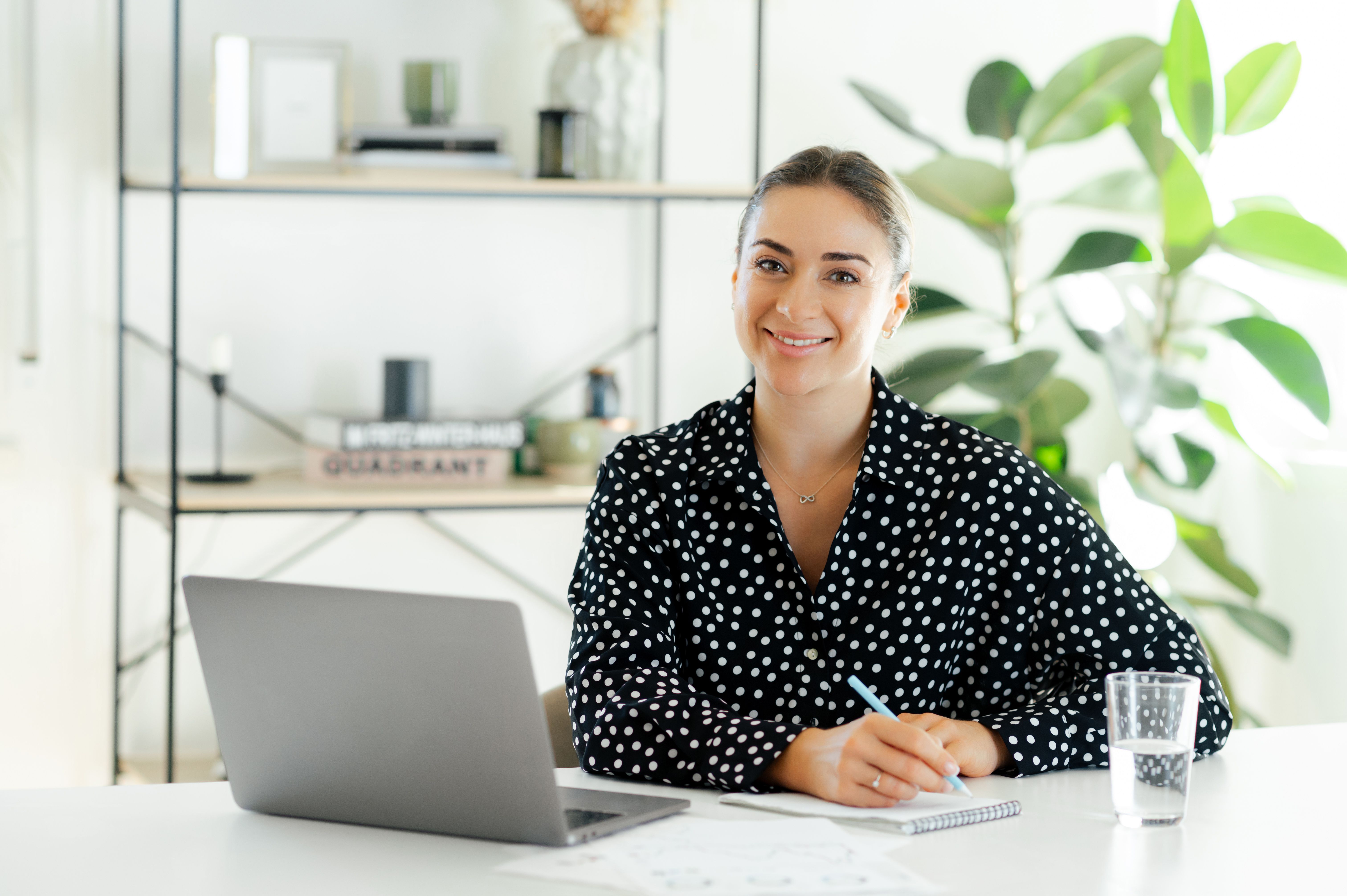 Glückliche erfolgreiche Geschäftsfrau, Managerin oder Sekretärin, die mit Laptop am Schreibtisch sitzt, einen Stift in der Hand hält und in die Kamera schaut, sieht in einem modernen Büro selbstbewusst aus und lächelt