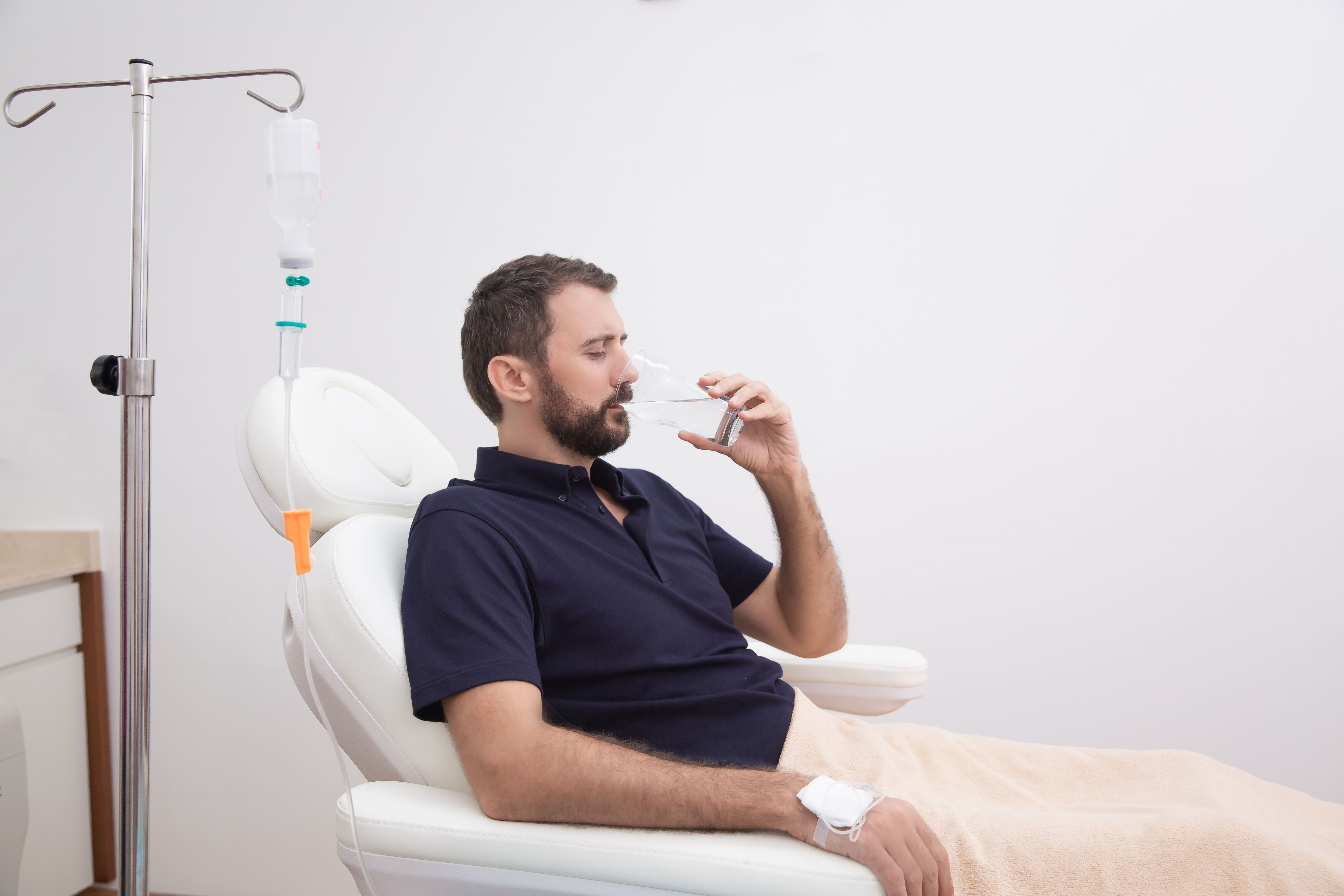 Sick, dehydrated or hangover patient man receiving vitamin IV infusion drip and drinking glass of water in hospital or beauty salon. Healthcare, wellness and medicine concept