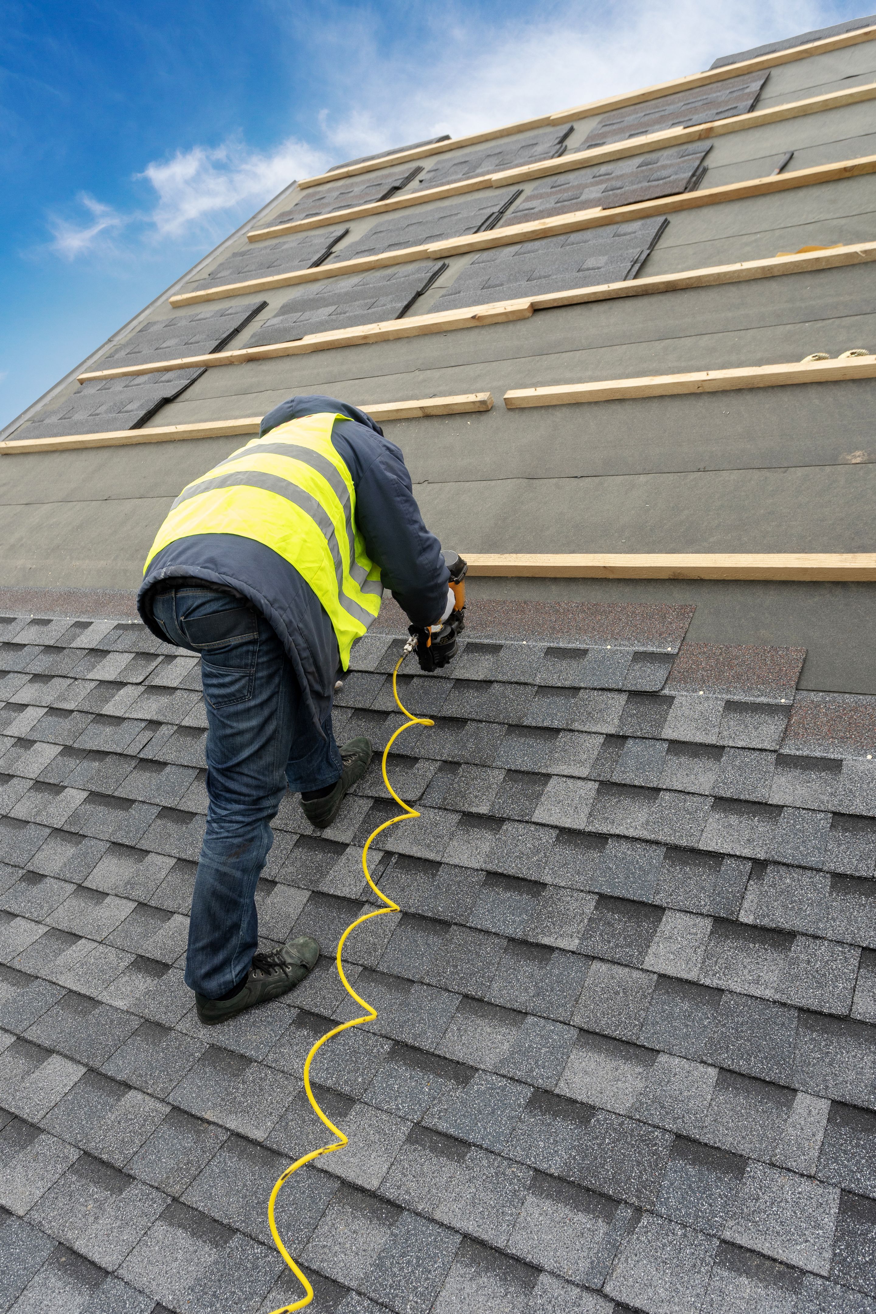 Vertical photo of unrecognizable roofer worker in uniform work wear using air or pneumatic nail gun and installing asphalt or bitumen tile on top of the roof under construction house Vertical photo of unrecognizable roofer worker in uniform work wear using air or pneumatic nail gun and installing asphalt or bitumen tile on top of the roof under construction house