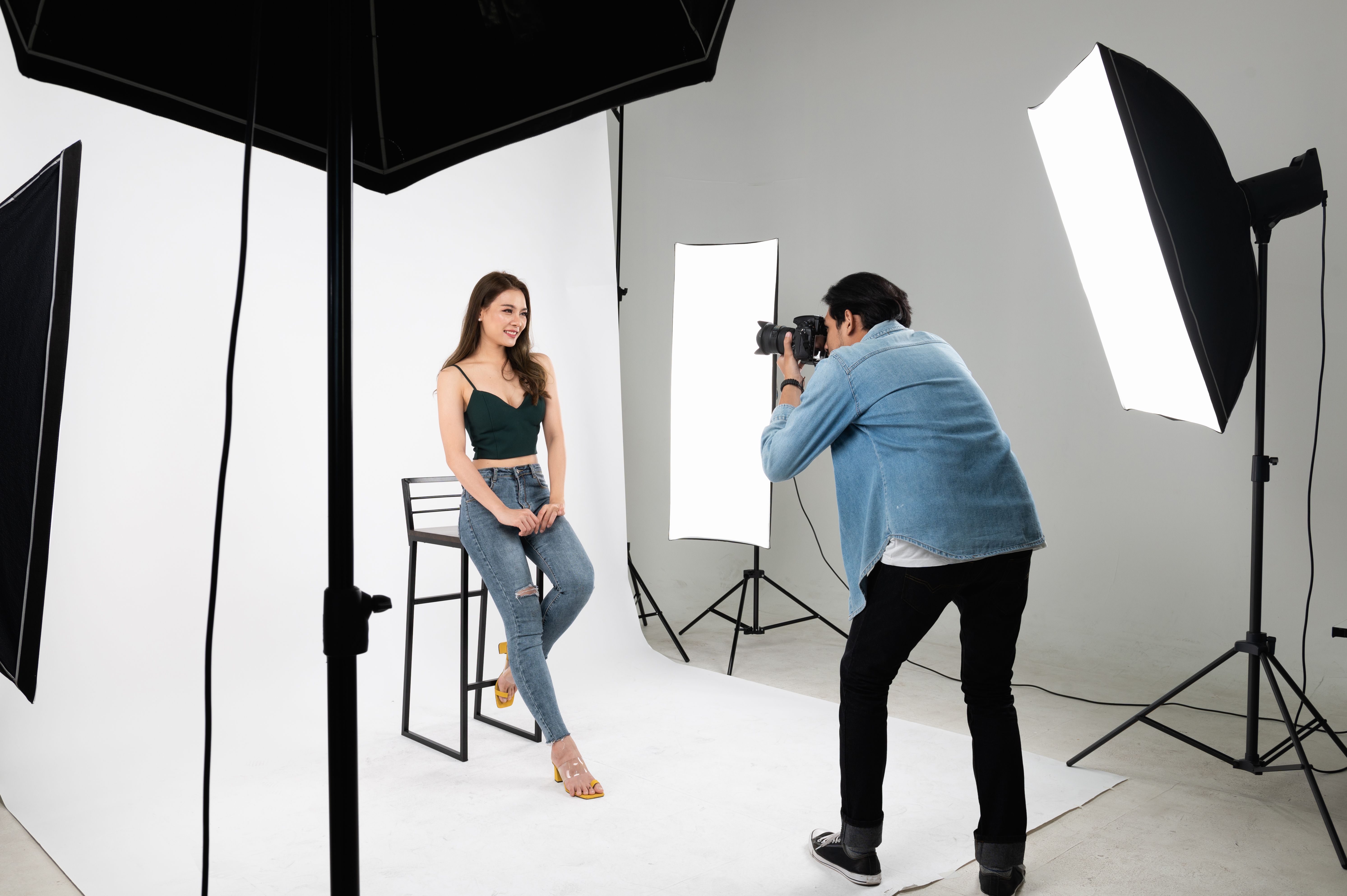Professional male photographer taking photo of Asian woman in indoor studio with professional set of photographic equipment.